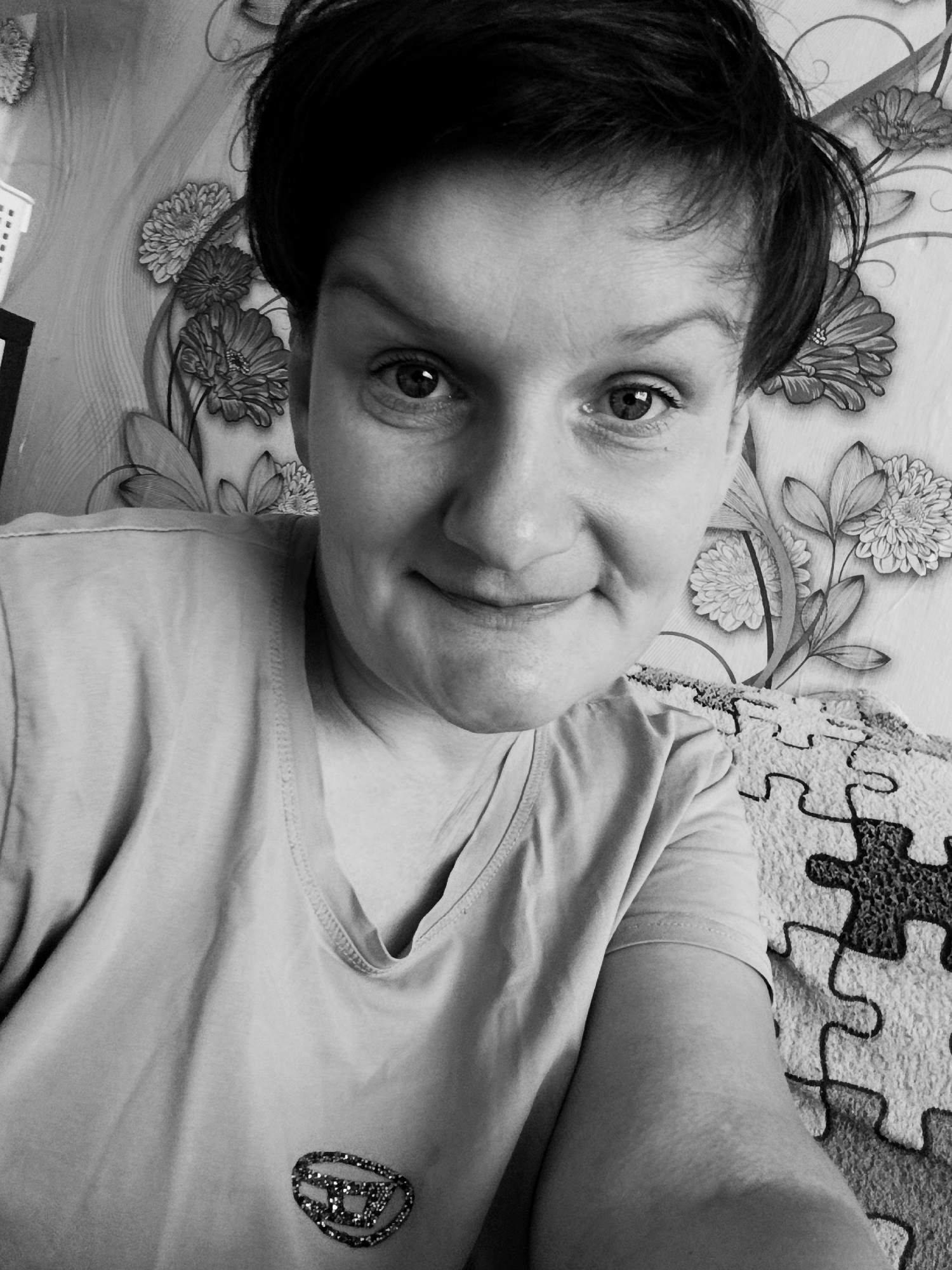 A black and white, close-up, selfie-style photograph shows a person with short, dark hair, looking directly at the camera. The person's face is partially obscured by the framing of the image, but their eyes, nose, and mouth are visible. They appear to be smiling slightly. The background consists of a floral-patterned wallpaper. The person is wearing a t-shirt with a decorative emblem on the chest. To the right of the person, a textured surface resembling a puzzle pattern is visible. The image quality is somewhat grainy. The location is not specified in the provided information.