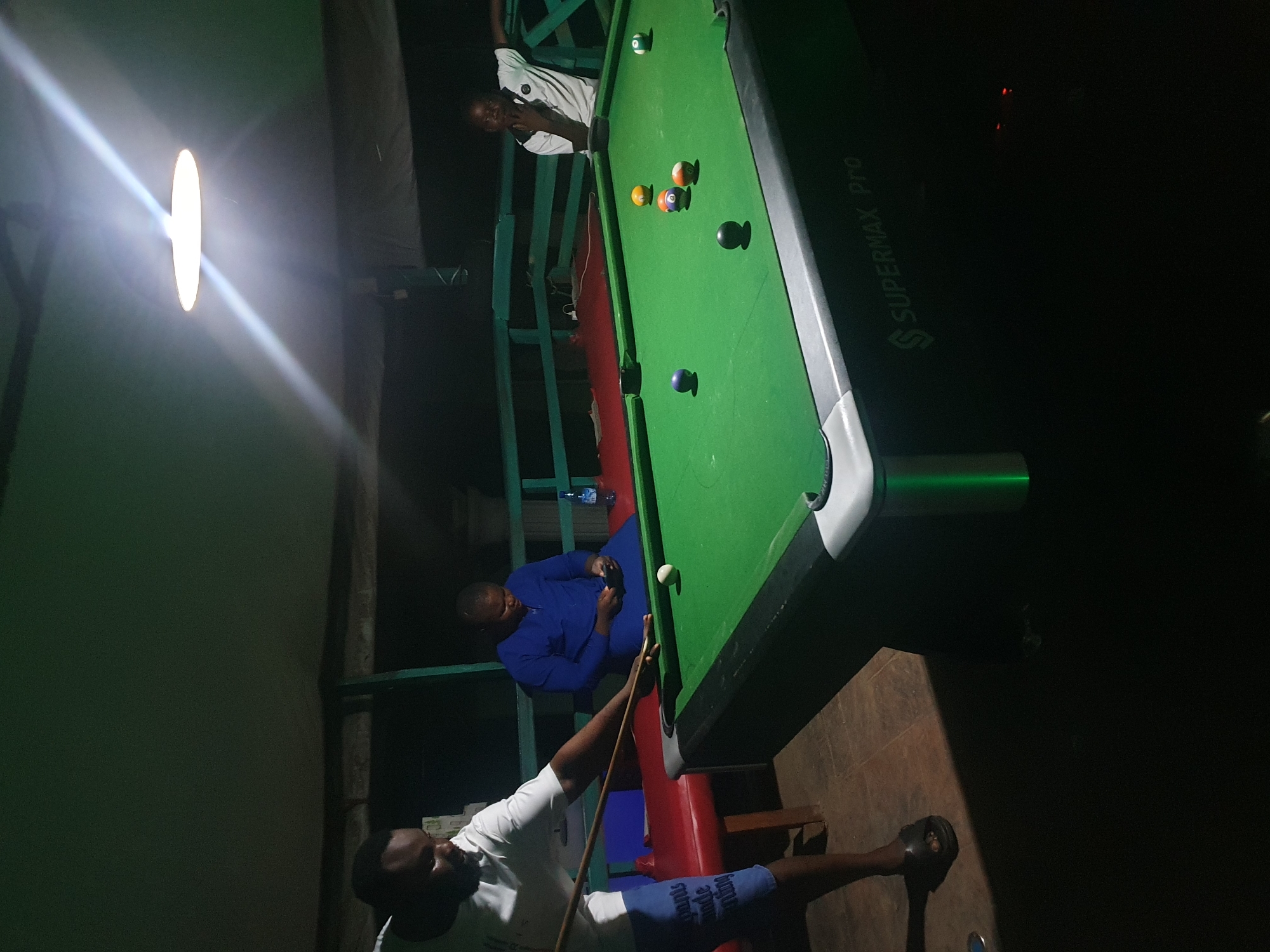 The image depicts an indoor pool hall in Lokoja, Nigeria, at night. A man is poised to take a shot on a green billiard table, holding a cue stick. Another person sits in the background wearing a blue garment, holding a phone. A third person, wearing a white shirt, leans on a railing. Several billiard balls are scattered on the table. A bright overhead light illuminates the scene. The environment appears to be an open-air structure, possibly a bar or recreational area, with a dark, textured floor.