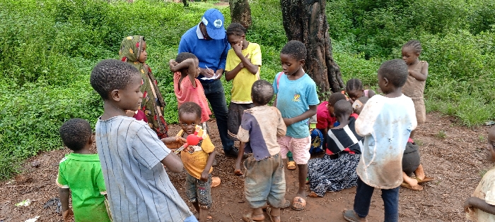 This image depicts an outdoor gathering in Ngalula, Democratic Republic of the Congo. One adult male and approximately 15-20 children are present. The adult male, positioned center-left, wears a blue long-sleeved shirt, dark pants, and a blue cap with a white circular logo. He is holding a white notepad and a pen, appearing to write. He faces towards a group of children.
The children, ranging in age from toddlers to pre-teens, are dressed in various simple, often worn and dirty, clothing items including t-shirts, shorts, and wraps. A boy in the immediate foreground, center-left, wears a light blue and white striped t-shirt and dark pants. To his right, another boy wears an orange t-shirt and holds a red and yellow object. A boy in a blue and beige t-shirt stands facing away from the camera in the central midground. A girl in a yellow t-shirt stands near the adult, with her hands crossed. Several other children are scattered around, observing the adult or interacting amongst themselves. Some children are partially obscured on the right side of the frame, where a group is seated or crouching. A young girl wearing a patterned headscarf and dress is visible on the far left.
The environment consists of an unpaved, reddish-brown ground, which appears damp in areas. Dense green foliage forms the background, with the trunk of a large tree visible behind the central group of people on the right.