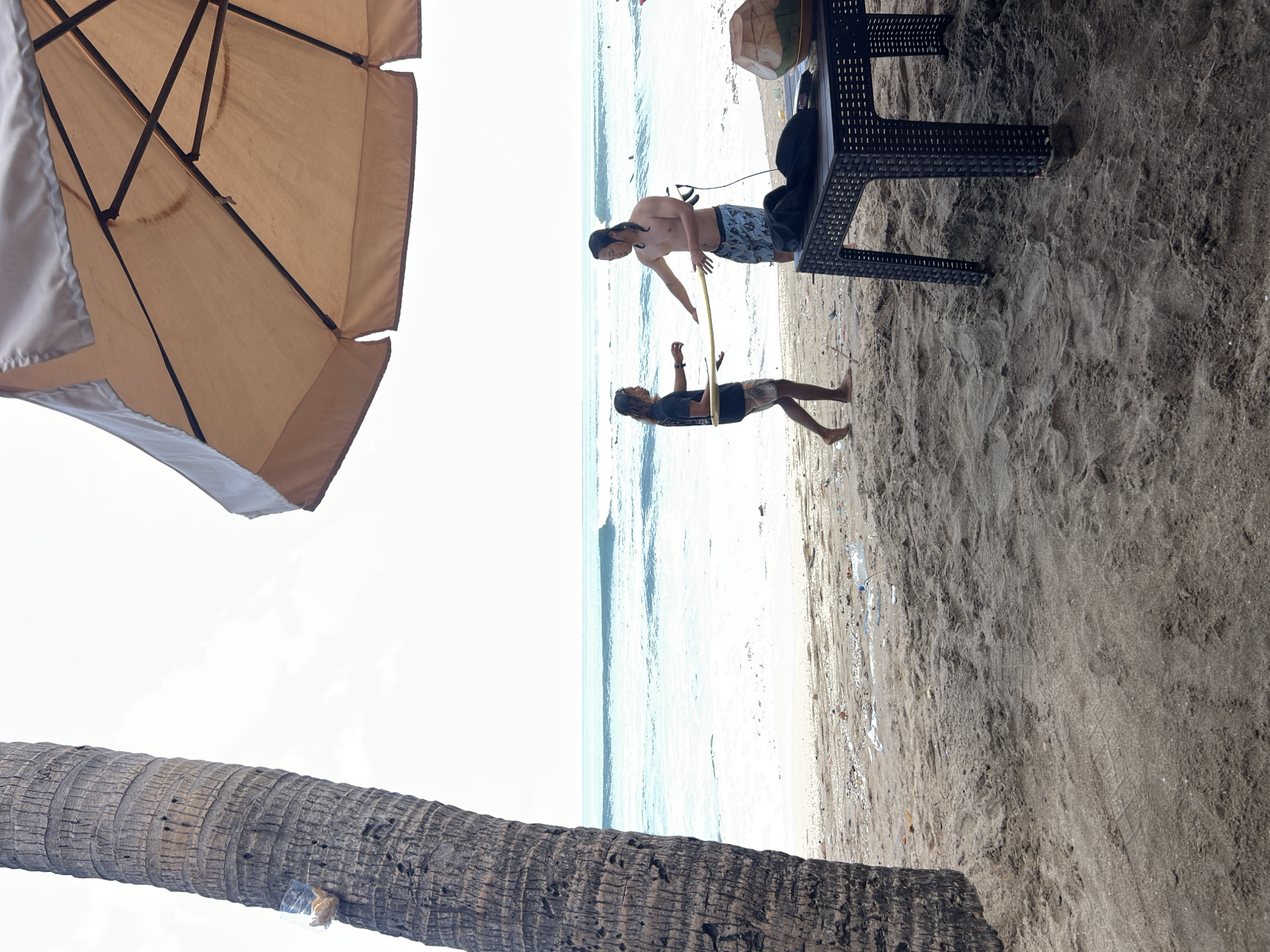 In Kuta, Indonesia, two men are on a sandy beach near the ocean. One man, wearing a dark t-shirt and shorts, stands on the left and holds a light-colored object. The other man, shirtless and wearing patterned shorts, stands on the right and also holds a light-colored object. They appear to be interacting with each other. In the background, the ocean has moderate waves, and the sky is bright and overcast. In the foreground, a large palm tree trunk is on the left, and a dark, woven table with a coconut on it is on the right. The beach is scattered with some debris. An umbrella shades the table and the area around it.