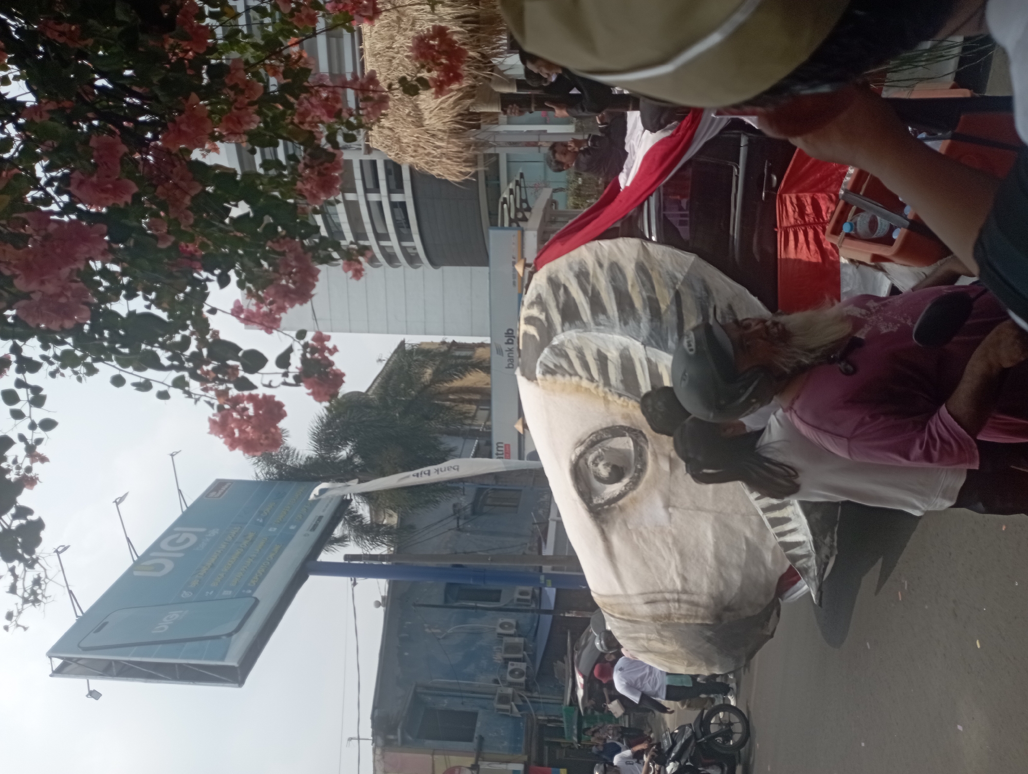 The image depicts a street scene in Purwakarta, Indonesia. In the foreground, a large, partially constructed float, resembling a fish or dragon head with a prominent eye and teeth, is visible. It is adorned with a red and white cloth, possibly the Indonesian flag. Several people are gathered around the float, some appearing to be constructing or preparing it. In the background, buildings line the street, including a blue-painted structure with visible air conditioning units and a modern, multi-story building. A large billboard advertising "DIGI" and "bank bjb" is prominently displayed overhead. The sky is overcast. The overall atmosphere suggests a street event or parade preparation.