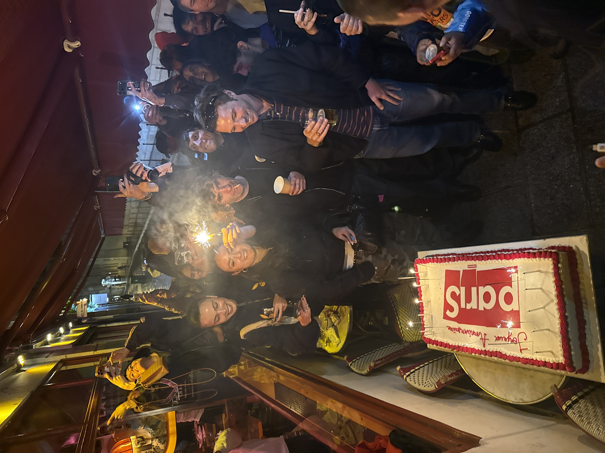 A group of approximately 15 people are gathered in an outdoor setting, likely in Paris, France, as indicated by the cake's inscription "Paris". The group is celebrating an event, possibly a birthday, as suggested by the cake decorated with sparklers and candles, and the inscription "Joyeux Anniversaire" (Happy Birthday). The cake is a rectangular shape with white frosting, red borders, and the word "Paris" written in a stylized font resembling the "Supreme" logo.
Many individuals in the group are holding up their phones, presumably taking photos or recording videos of the celebration. Some are holding drinks and/or sparklers. The environment appears to be a restaurant or bar with a canopy structure visible above. The lighting is somewhat dim, supplemented by the flashes of the phone cameras and the light from the sparklers. Some people are smiling and engaging with the camera, while others are focused on the cake and the celebration. The overall atmosphere appears celebratory and joyful.