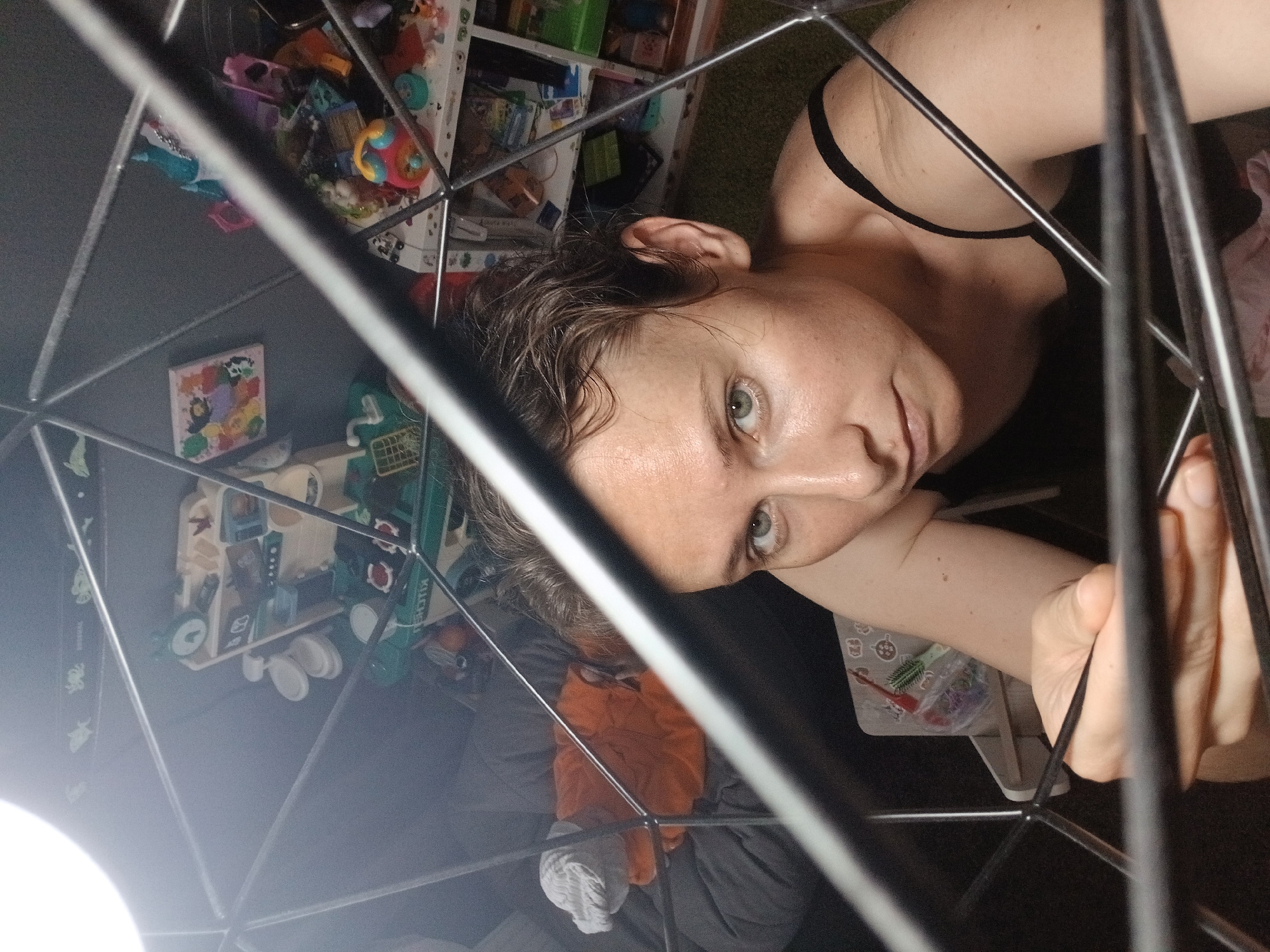 The image shows a medium shot of a woman, positioned slightly off-center to the right, looking directly at the camera. She has short, dark, wet-looking hair and light green eyes. Her skin appears to be fair with some texture visible. She is wearing a black tank top. The woman is partially obscured by a dark, metallic geodesic frame, which dominates the foreground and creates a sense of depth.
The background is out of focus but reveals a cluttered environment that appears to be a child's play area or a room with many toys. To the left, a grey sofa with an orange blanket and a white cloth item is visible. Further back and to the right, shelves are filled with various colorful toys, including what appears to be a toy kitchen set with small white plates. The overall impression is of an indoor setting, possibly a home.
There is no discernible information in the image that would suggest a location in Yekaterinburg, Russia, or any other specific city. The image is a close-up, personal shot focused on the woman and her immediate surroundings, with no external environmental cues.