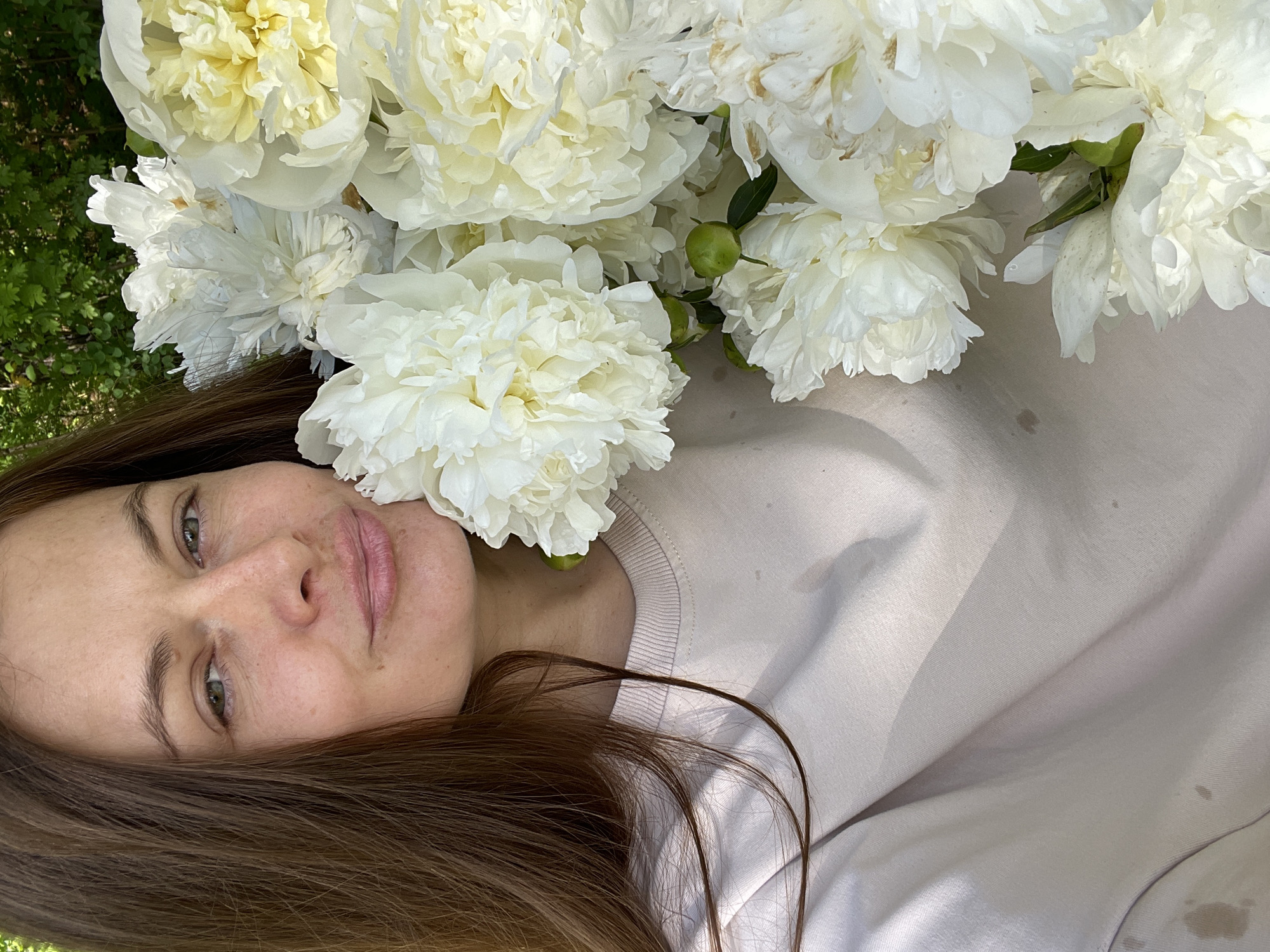 A woman with long brown hair and fair skin, wearing a light pink sweatshirt, is holding a large bouquet of white peonies. She is looking towards the camera with a slight smile. The background is blurred, but appears to be an outdoor setting with green foliage, suggesting a garden or park. The image is cropped to focus on the woman's face and the flowers, with only a portion of her upper body visible. There are no identifiable landmarks or signs indicating a specific location within the city of Zarechny, Russia.