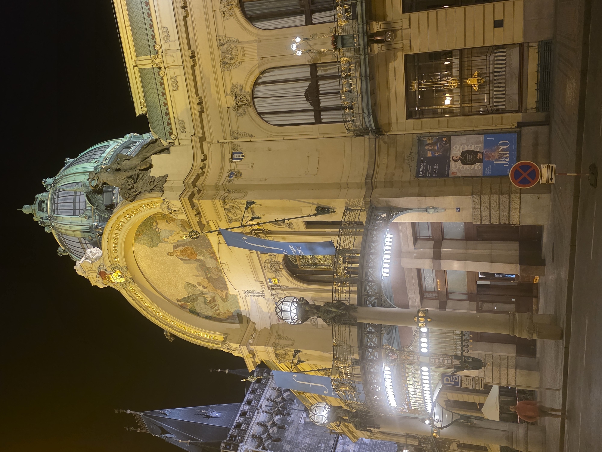 Here is a detailed description of the image:
**Scene:** The image captures the Municipal House (Obecní dům) in Prague, Czech Republic at night.
**Architecture:** The Municipal House is a striking example of Art Nouveau architecture. Notable features include:
* A large dome with sculptures and a mosaic.
* Ornate architectural details, including arched windows, decorative carvings, and metalwork.
* A façade with a fresco and blue flags.
**Environment:** The scene is illuminated by streetlights and lighting on the building, with the sky mostly dark. A street runs alongside the building.
**Objects:**
* Street lamps
* A parking sign and a "No Parking" sign are visible.
* Posters are displayed on the building, with one showing "BELTRACCHI" and "Jaro".
**People:**
* A pedestrian in a red jacket is walking on the street.