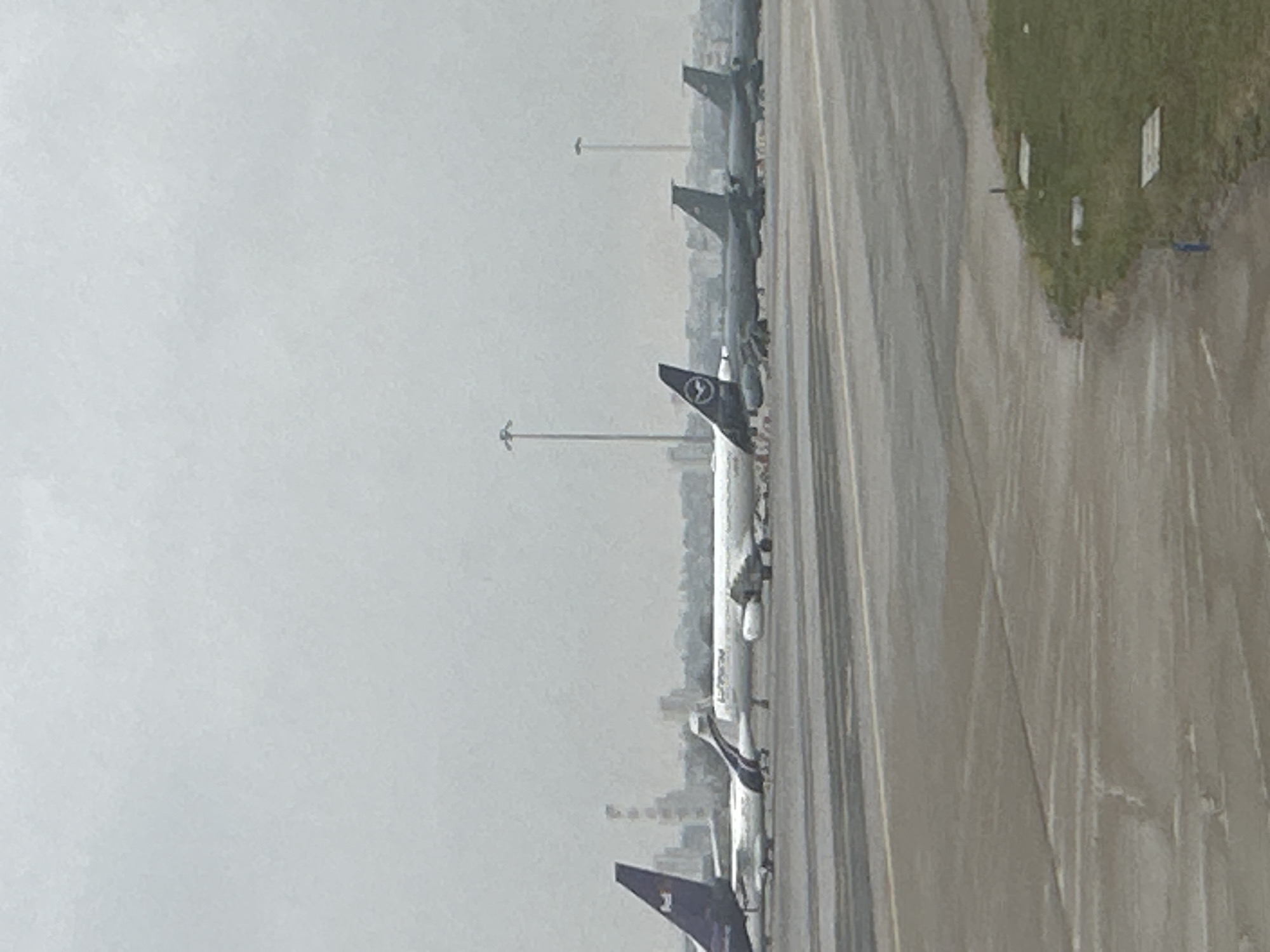 The image shows a lineup of Lufthansa airplanes at Sofia Airport in Sofia, Bulgaria, on a runway. The lighting is overcast. There are taxiway lights visible in the middle ground. There is grass on the shoulder of the runway. Buildings are visible in the background.