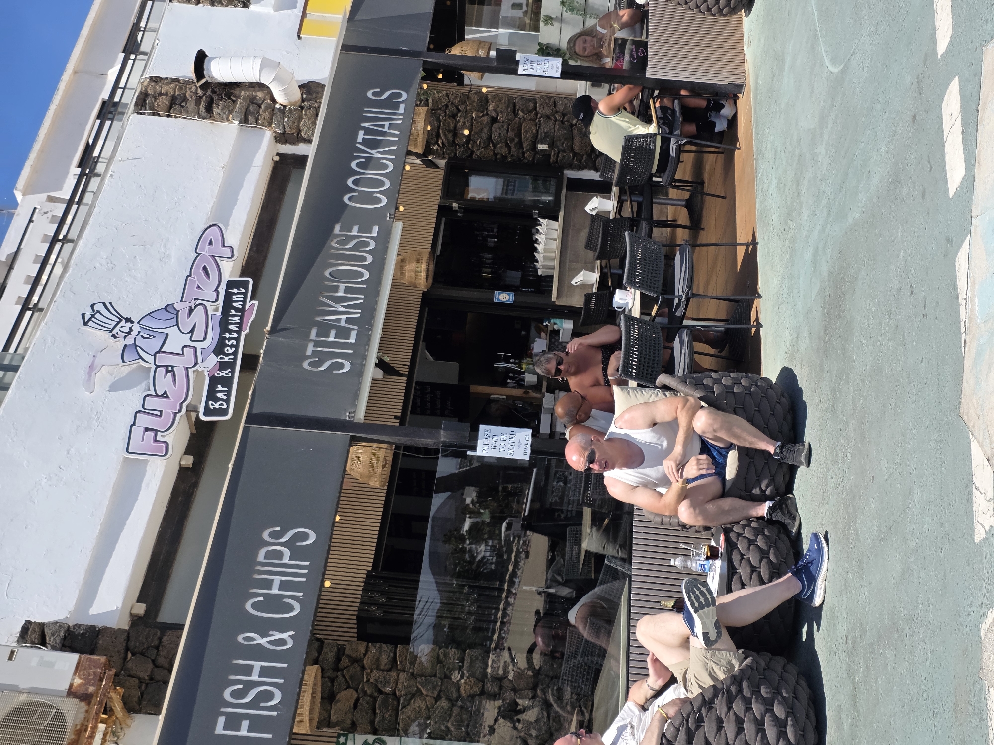 The image depicts an outdoor seating area of a restaurant and bar named "FUEL STOP" located in Teguise, Spain. The signage above the establishment advertises "FISH & CHIPS" and "STEAKHOUSE COCKTAILS." Several individuals are seated at tables and on comfortable outdoor chairs. Two men in the foreground are relaxed, with one wearing a white tank top and shorts, and the other wearing a white t-shirt and shorts. A bottle of water and a drink with ice are visible on a low table between them. In the background, other patrons are seated at tables, with some interacting and others looking at their phones. The setting appears to be a sunny day, with the restaurant's facade and outdoor dining area clearly visible.