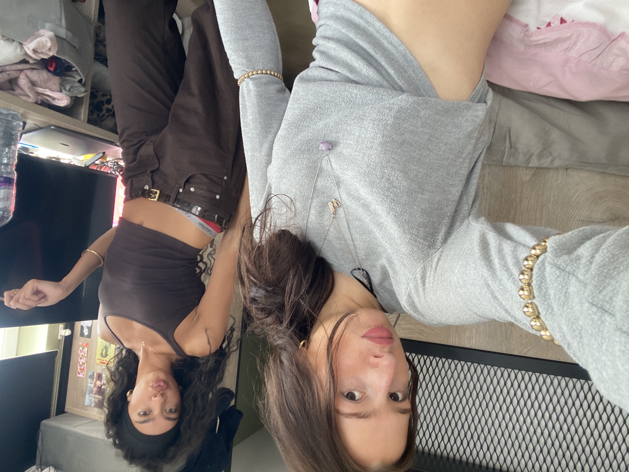 Two women are visible in the image, which appears to have been taken with the camera oriented upside down. One woman is wearing a brown tank top, brown pants with a visible Calvin Klein underwear waistband, and a gold bracelet. The other woman is wearing a gray long-sleeved shirt and a gold bracelet. The background includes a television, a shelf with items, a framed picture, a bed with pink and gray bedding, and a black metal mesh surface. There is no indication of a London location within the image.