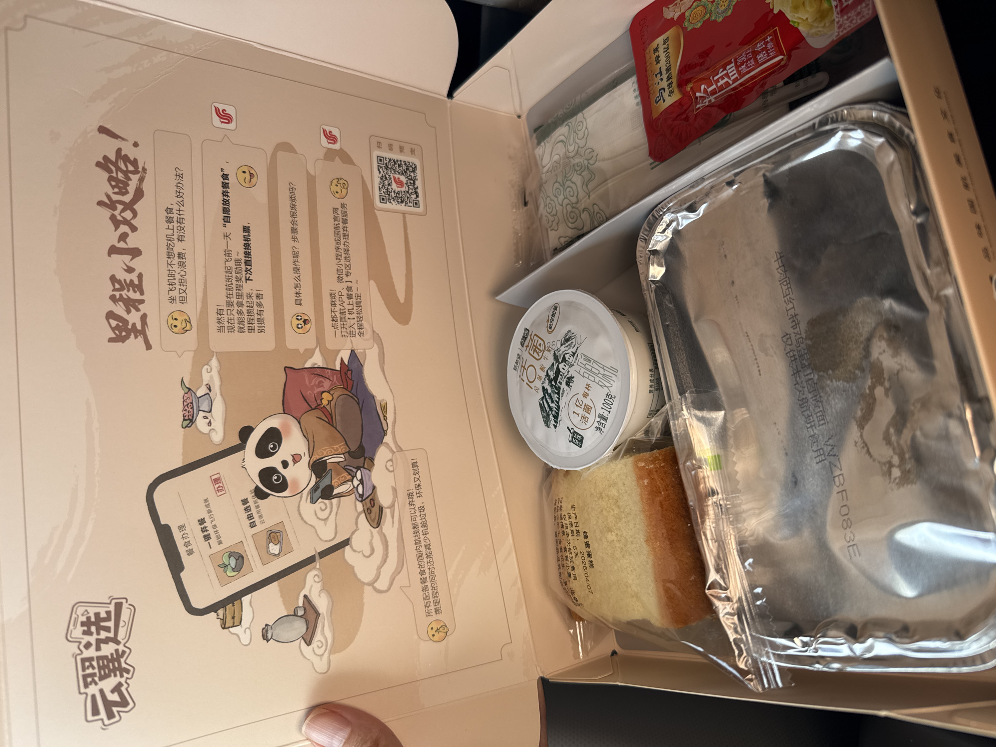 Here is a forensic description of the image:
**Scene:** An open in-flight meal box on what appears to be an airplane.
**Objects:**
* The open meal box has text and cartoon graphics printed inside its lid, describing a "mileage strategy" and food waste reduction. The text is in Chinese.
* Inside the box are an aluminum foil meal container, a packaged piece of cake, a small container of yogurt, a packet of what is presumably Chinese pickled vegetables (brand name “Wu Jiang”), a packet of utensils, and a red packet with the text 牛肉西红柿鸡蛋打卤板面 "beef tomato egg stewed noodle" printed on it.
* The aluminum container has "牛肉西红柿鸡蛋打卤板面 WZBF083E" printed on it, also.
**Actions/Interactions:**
* The box is likely being held by someone, as a fingertip is visible in the bottom-left corner.
**Location:** It is impossible to determine whether the scene is in Tashkent, Uzbekistan from the given image.
