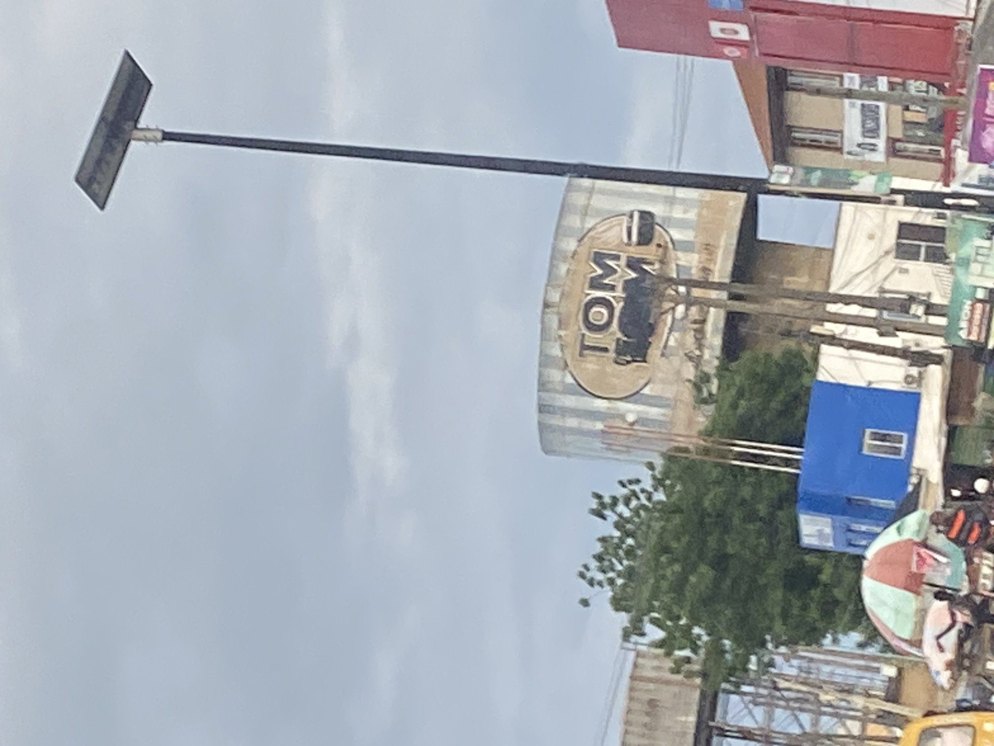 In Ikorodu, Nigeria, an outdoor scene unfolds under a cloudy sky. A tall, thin lamppost with a solar panel at its apex dominates the right side of the frame. To the left, a large, cylindrical water tank with a weathered sign reading "TOM TOM" is visible. The sign features a circular emblem with text and an image of what appears to be a bowl.
At the base of the water tank, a cluster of structures creates a bustling atmosphere. A bright blue, cubical building with a white-framed window stands out. In front of it, a colorful umbrella with segments of red, white, and green provides shade over a market stall. Several people are present, some partially obscured. A yellow vehicle is visible on the far left. Further to the right, a brown-roofed building with multiple windows is situated, and a prominent red building with white signage extends into the frame. Other signs with various text and graphics are scattered throughout the scene, suggesting a commercial or market area. The overall impression is one of a lively, urban environment.