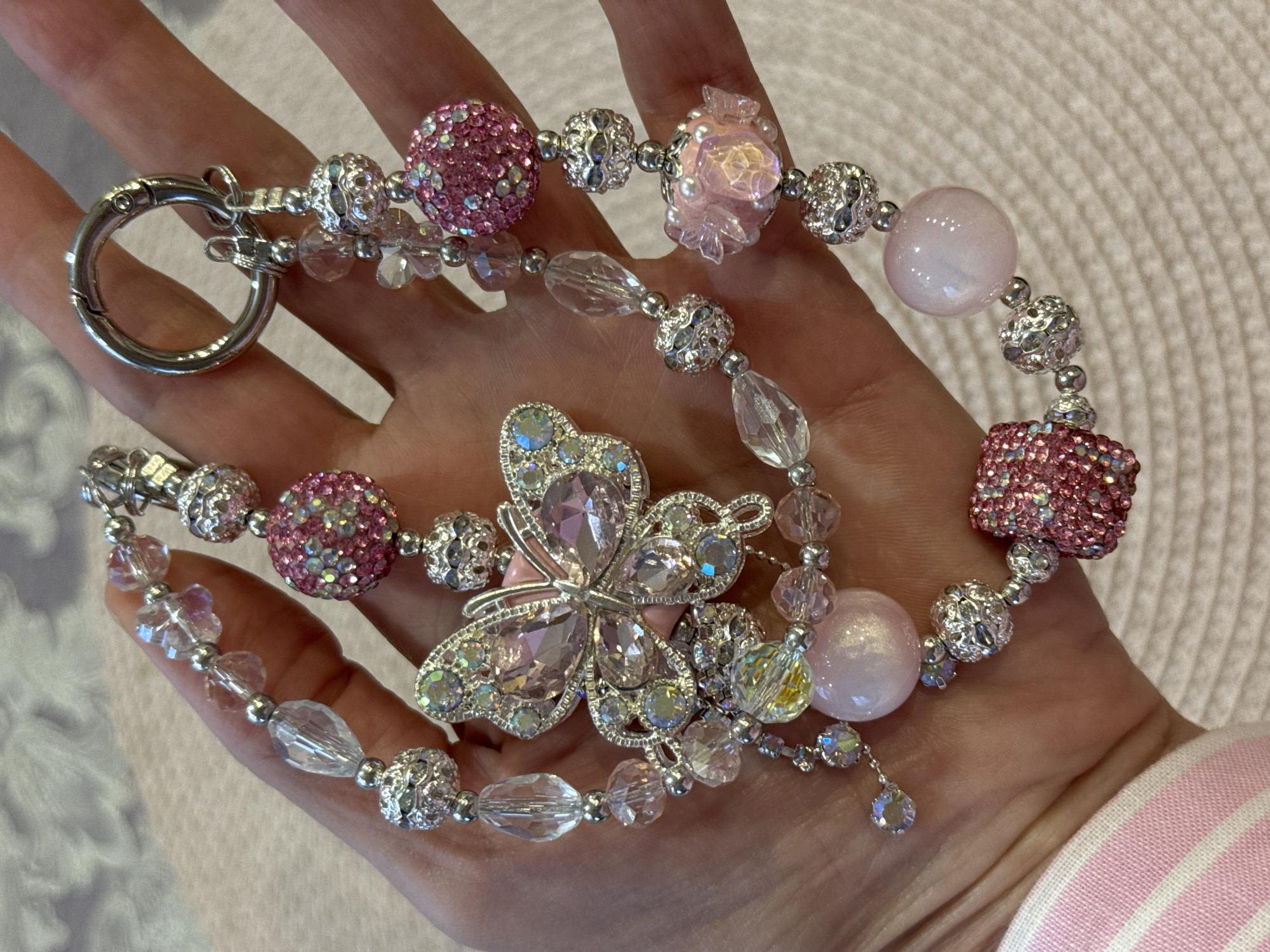 An ornate, bejeweled charm dangles from a person's hand. The charm features a prominent butterfly embellished with clear and pink rhinestones, surrounded by a cascade of various beads. These beads include multifaceted clear crystals, smooth pale pink opaque spheres, and intricately textured silver-toned beads. Some sections of the charm are densely encrusted with small pink and iridescent rhinestones, forming spherical or rectangular shapes. A silver-toned clasp is attached to the top of the charm, suggesting it's designed to be attached to a bag or other accessory. The background is a pale, textured surface. There is no indication of the location being in Unknown, Russia.
