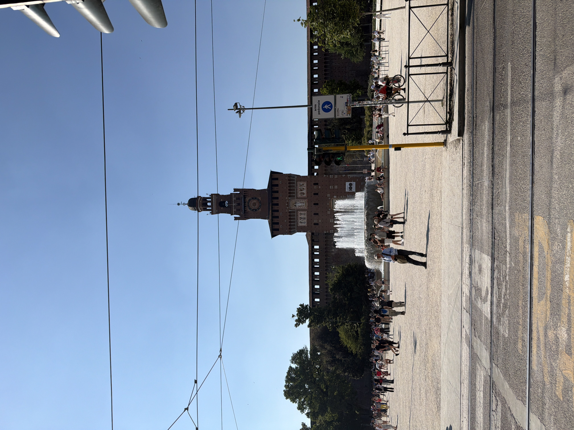 The image is an outdoor shot of Sforza Castle in Milan, Italy, taken during the day. The castle's tower rises prominently against a clear, blue sky. Overhead wires are visible in the foreground. A fountain is running near the castle, and a crowd of people is gathered in front of it. A street with tram rails and markings runs alongside the plaza where the people are standing. The shot is taken from a low angle and is oriented vertically.