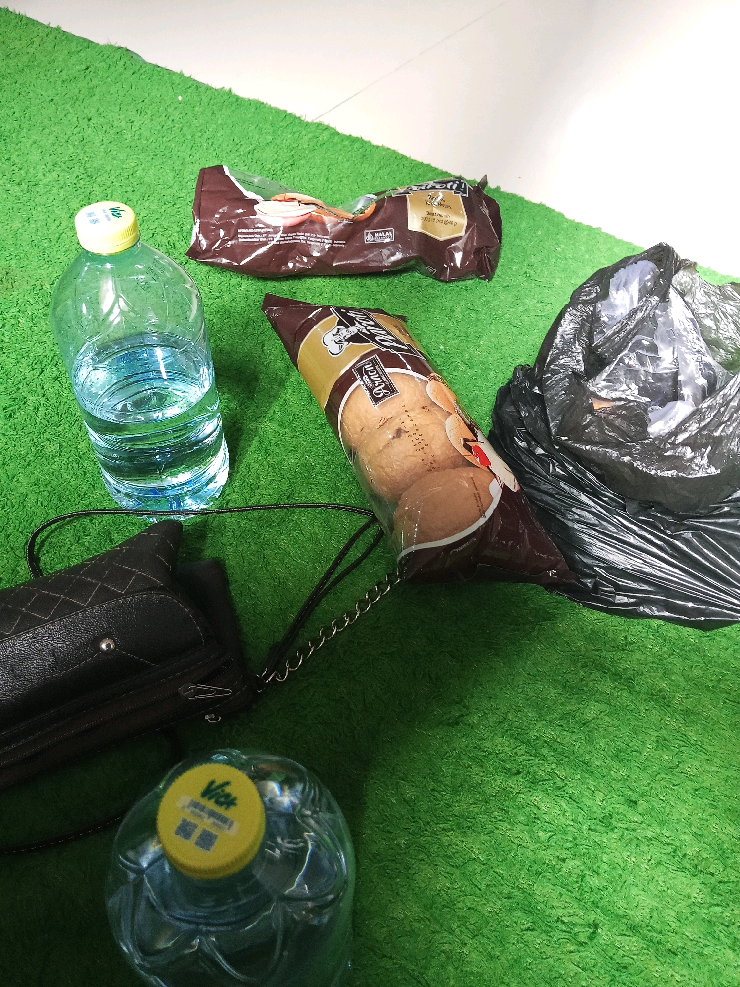 A high-angle, close-up shot captures several items scattered on a vibrant green, shag carpet. In the foreground, a clear plastic bottle of water with a yellow "Velt" brand cap sits to the left. Adjacent to it and slightly to the right is a dark brown, textured handbag with a silver zipper and chain strap. To the upper right of the handbag is a black plastic trash bag, partially filled and crumpled. Further to the right and slightly above the trash bag is a large, crinkly plastic package containing several golden-brown buns or bread rolls. To the left of this bun package, and partially overlapping it, is a smaller, dark brown foil package of cookies, featuring a "HALAL" sticker. The background is a bright, plain white surface. The overall impression is a collection of everyday items, possibly indicating a break or a planned outing, given the presence of snacks and drinks. Based on the context of the "Velt" water brand, the location is likely to be Kediri City, Indonesia, where this brand is commonly found.