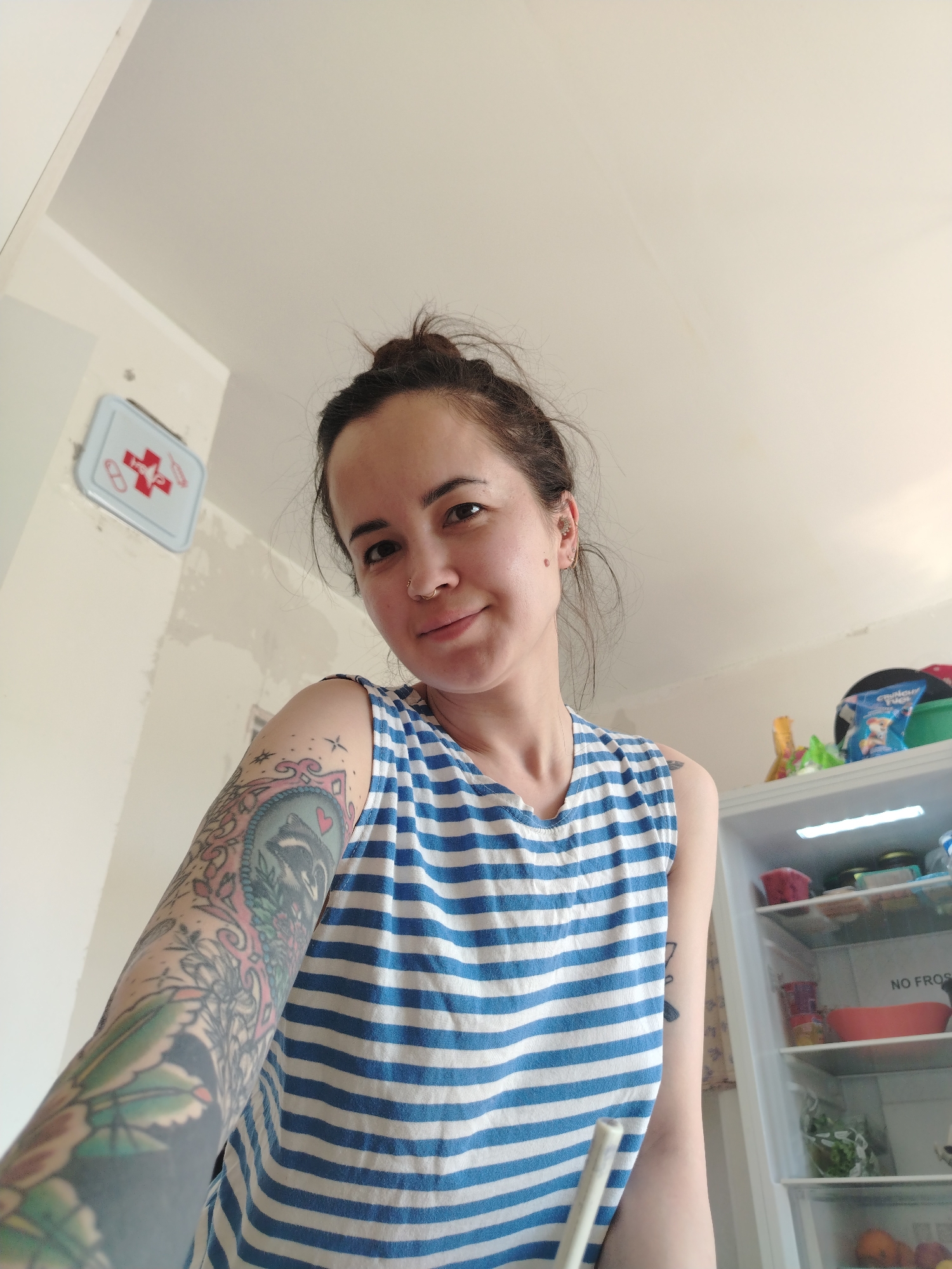 A woman stands indoors, likely in a kitchen. She is fair-skinned with dark hair styled in a messy bun. She has a nose ring and a mole on her right cheek. Her left arm is heavily tattooed. She wears a blue and white striped tank top. Behind her, a white refrigerator is partially visible, stocked with food items. To her left, a wall-mounted first aid kit is visible on a partially painted wall. The location is likely a domestic setting in Noviy, Russia.