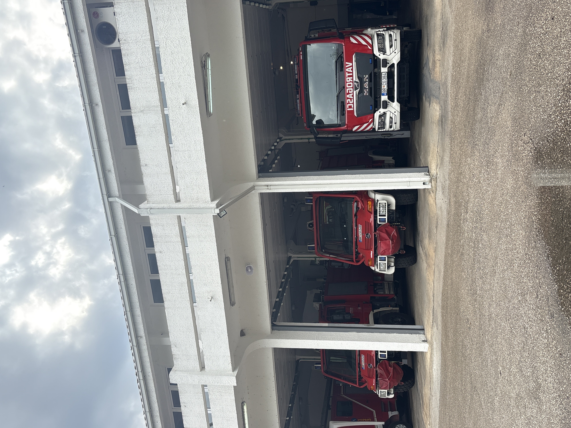 The image, originally rotated 90 degrees clockwise, captures the front of a fire station in Bar, Montenegro, under a cloudy daytime sky. Three red fire trucks are parked inside individual bays of a multi-bay, white building.
The building has a textured white facade with several dark-paned windows on the upper level and a gutter system along its roofline. The ground in front of the bays is paved with asphalt or concrete.
In the top bay, a large, modern red fire truck is visible. Its front bumper and grille are white with red striped detailing. A "MAN" logo is visible on the grille, and its license plate reads "BR C6384". The word "VATROGASCI" (Firefighters) is reflected on the windshield banner.
The middle bay contains a smaller red fire truck. Its front features white sections, and its headlights are covered with red fabric. The license plate displayed is "BR C0148".
The bottom bay houses another smaller red fire truck, similar to the one in the middle bay, also with red headlight covers and the license plate "BR C0148".
The sky is overcast, suggesting a cloudy day. No people or active events are present; the fire trucks are stationary. The "BR" on the license plates indicates the city of Bar, aligning with the provided location context.