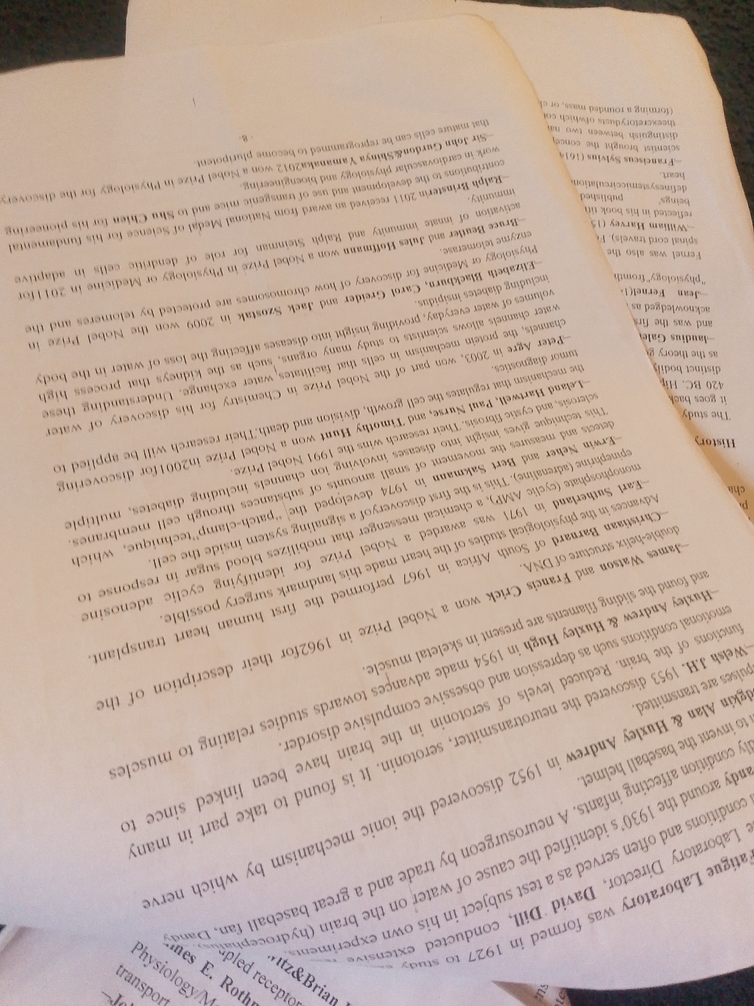 The image shows a close-up, angled view of several pages of a book with text printed in English. The pages are slightly creased and worn, suggesting they are from an older publication. The text is printed in black ink on off-white paper. The book appears to be a collection of biographical or historical entries, as indicated by names like "Sir John Gurdon," "Ralph Steinman," "Elizabeth Blackburn," and "James Watson." The entries mention scientific discoveries and Nobel Prizes, such as for "telomeres," "dendritic cells," and "DNA." The lighting in the image is somewhat uneven, with some areas brighter than others, and there are shadows cast by the book's pages. The background is out of focus and appears to be a neutral surface. There is no identifiable information suggesting a location in Jalingo, Nigeria, or any specific environmental details related to a city.