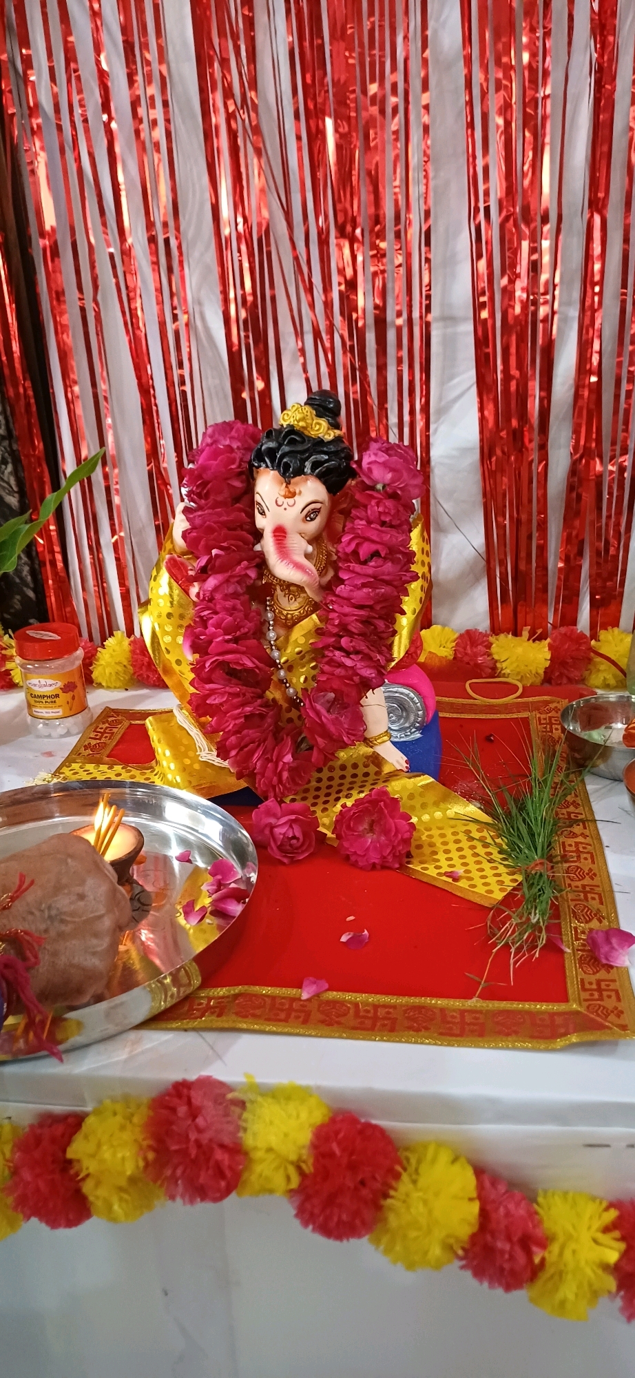 A vertical, eye-level, full shot shows a decorated Ganesh idol, likely for a religious festival, arranged on a surface. The background is adorned with alternating red and white shiny vertical streamers, suggesting a festive backdrop. In the foreground, a silver plate holds a lit candle and a small piece of something that appears to be food or an offering, possibly covered by a hand. To the left of the plate, a small orange and white container labeled "CAMPHOR 100% PURE" is visible, along with yellow and red pom-pom decorations. The Ganesh idol itself is adorned with a vibrant pink garland of flowers and a yellow and gold patterned cloth. It is placed on a red mat with a golden swastika design border. A small bunch of green grass is also placed on the mat. The scene suggests an indoor setting, possibly a home or a small shrine, in Ajmer, India.