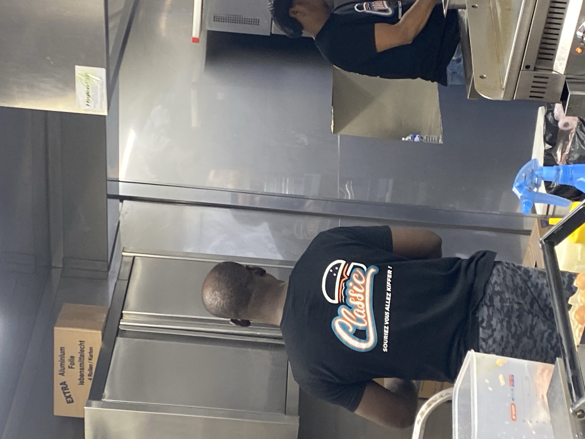 A man with a shaved head is wearing a black t-shirt with a colorful logo that says "Classic! Souriez Vous Allez Kiffer!" and gray camouflage pants. He is standing in what appears to be a commercial kitchen, possibly in Schiltigheim, France. There is a box labeled "EXTRA Aluminium Folie Lebensmittelrecht 4 Rollen / Karton" and an industrial-looking refrigerator with a label that says "Hygienistar." There is another man wearing a black shirt with an American flag on it, possibly working on a grill. A blue spray bottle is visible near a stainless steel surface. The background includes stainless steel appliances and surfaces.