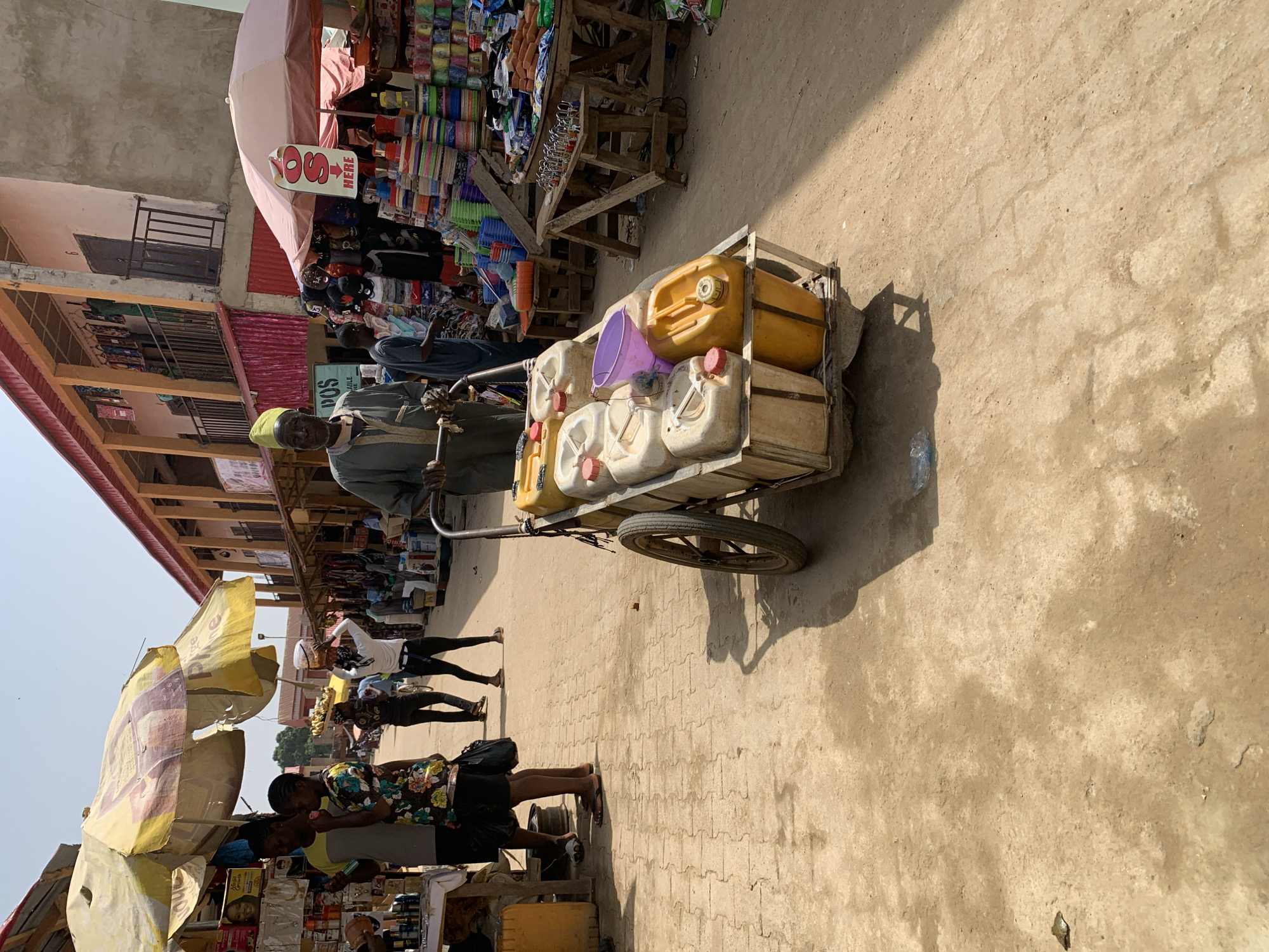 The image captures a bustling street market scene in Kafanchan B, Nigeria, under bright sunlight. The central subject is a man, wearing a light-colored shirt and trousers with a bright green cap, pushing a two-wheeled wooden handcart. The cart is heavily loaded with multiple plastic jerrycans, mostly white and yellow, along with a purple bucket and other containers, indicating the transport of liquids such as water or fuel.
The ground is a mix of packed earth and interlocking paving stones, with distinct shadows cast by the sun, suggesting it is mid-morning or afternoon. On both sides of the street, market stalls are set up, covered by various umbrellas and canopies that provide shade. One large yellow umbrella with faded text is visible on the left.
The stalls are filled with a diverse array of goods. On the right, under a light pink canopy, stalls display numerous colorful plastic items like bowls and containers, as well as clothing. A white sign with red text "SOS HERE" and an arrow pointing is attached to this canopy, likely indicating a shop or service. Further down the street, buildings with concrete facades and balconies are visible.
Several other people are present in the market, walking, standing, and engaging with the stalls, contributing to the lively atmosphere. One person in a white top and dark trousers is seen near a stall, and another in a floral-patterned shirt is closer to the foreground. The scene depicts everyday commerce and activity in a vibrant outdoor market setting. A partial word "POS" is visible on the man's chest.