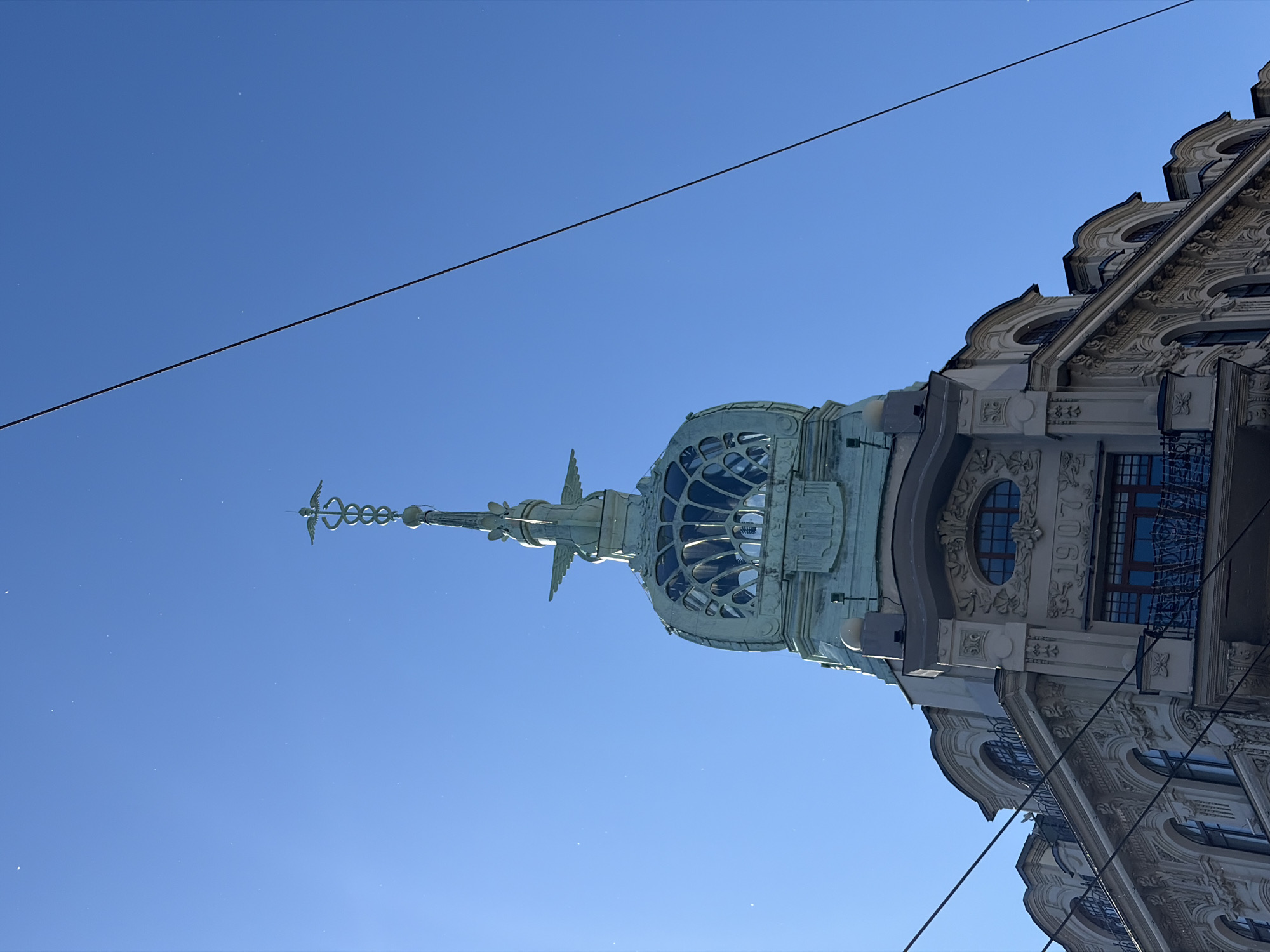 The image shows the top section of a building in Saint Petersburg, Russia, under a clear blue sky. The building features ornate architectural details, including intricate carvings and a domed roof with decorative elements. A prominent feature is the Caduceus symbol, with wings and a coiled serpent, atop the spire of the dome. The date "1907" is visible on the building's facade. Electrical wires cross the sky, diagonally bisecting the upper portion of the frame. The perspective is from a low angle, looking upwards at the building's upper stories and spire.