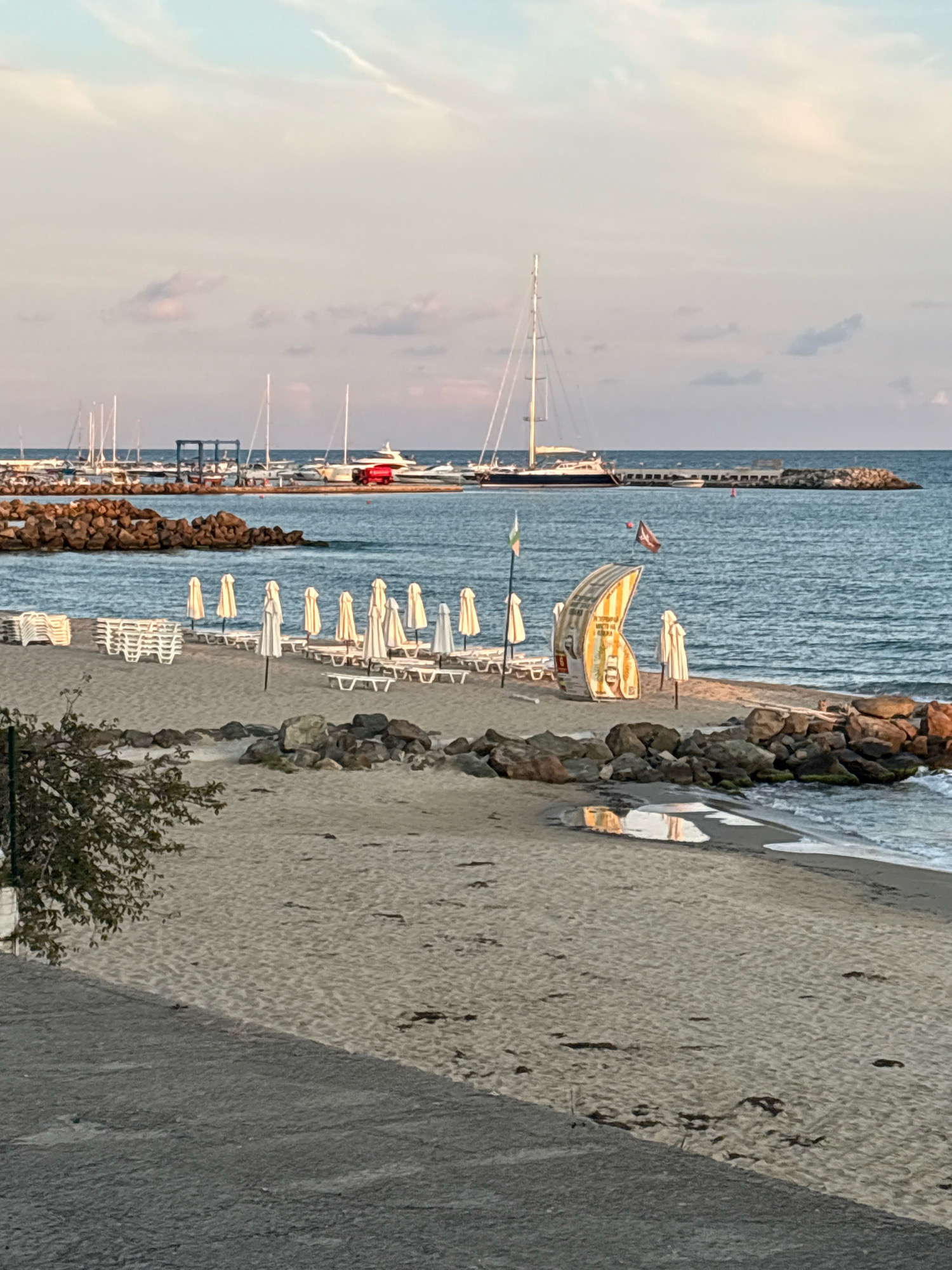 The image depicts a serene beach scene in Sveti Vlas, Bulgaria, likely during the late afternoon or early evening. In the foreground, a sandy beach stretches towards the gentle waves of the Black Sea. Numerous white beach chairs are neatly stacked on the sand, alongside several white beach umbrellas, some of which are open. A distinctive curved, yellow and white structure, possibly a lifeguard station or a beach amenity, stands prominently on the beach.
In the midground, the sea is calm, with a rocky breakwater extending into the water. Beyond the breakwater, a marina is visible, filled with various sailboats and yachts, their masts creating a delicate silhouette against the sky. A larger sailboat with a dark hull and tall masts is particularly noticeable. The sky is a soft pastel of blue and pink, with scattered clouds, indicating either dawn or dusk.
Further in the distance, a long pier or breakwater stretches across the water, and a red boat is moored near it. The overall atmosphere is tranquil and picturesque, capturing the essence of a seaside resort town. The image is taken from a slightly elevated perspective, suggesting it was captured from a balcony or a higher point overlooking the beach and marina.