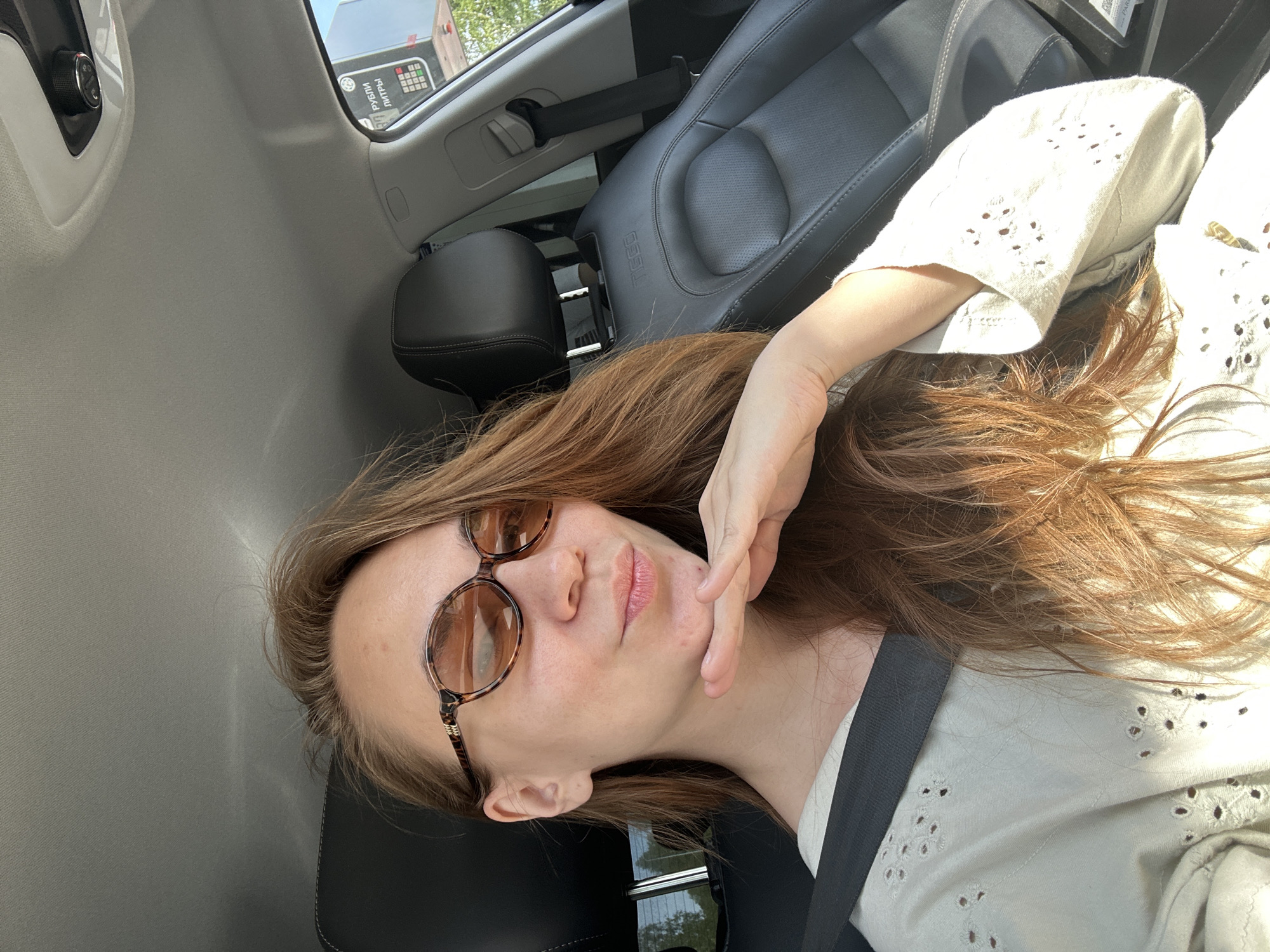 A woman is seated in the driver's seat of a car, wearing a seatbelt, a white shirt with a patterned cut-out design, and brown-tinted glasses. Her right hand is near her face. The interior of the car is visible, including the grey seats and car fixtures. Through the car window, part of a building with text written in Russian is visible. Based on the text, the location is inferred to be in Uva, Russia.