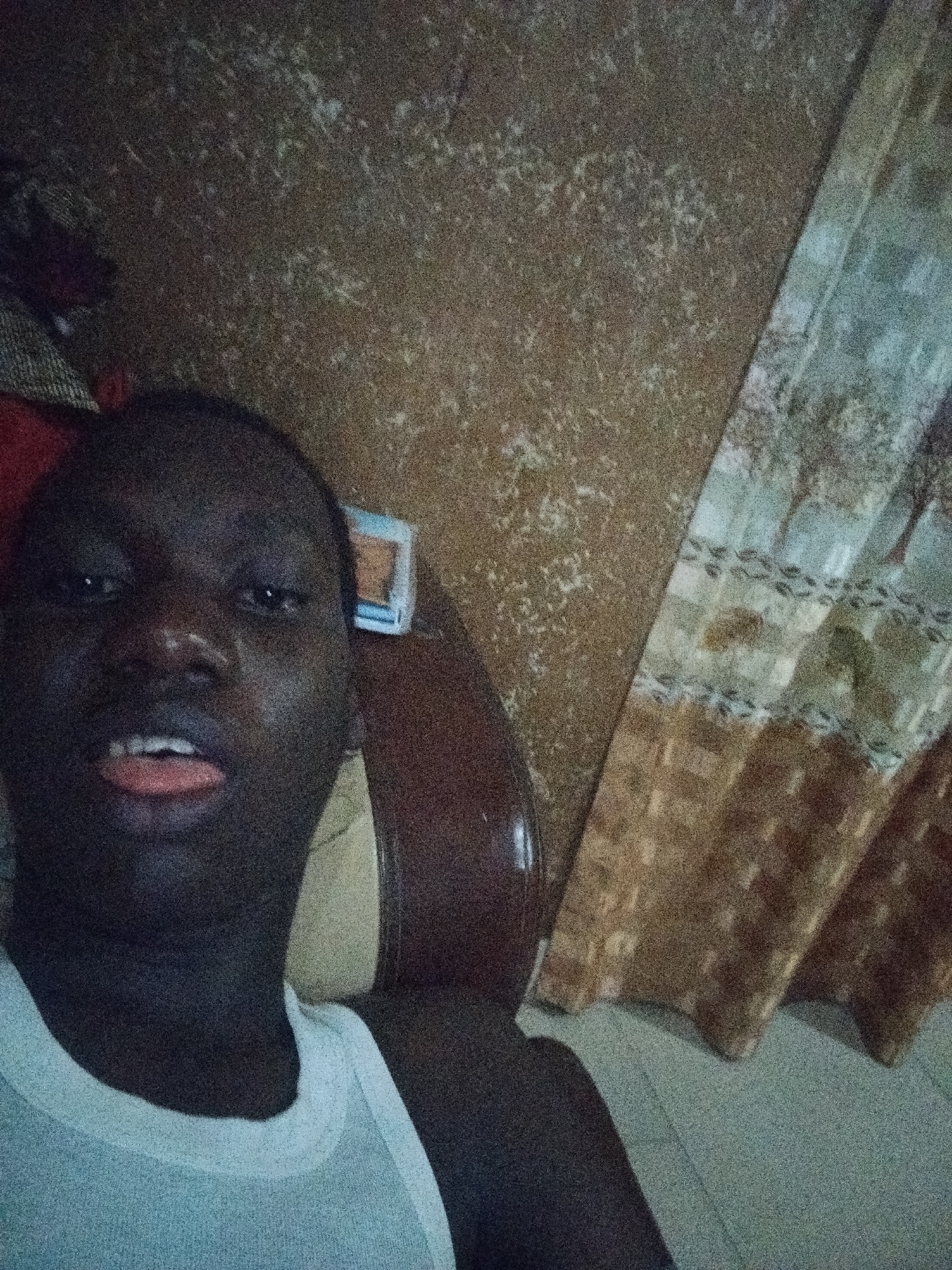 A dimly lit selfie of a dark-skinned man wearing a white tank top. He is looking directly at the camera with his tongue slightly protruding. Behind him is a brown table with a small blue and white object on it. The background consists of a textured wall and patterned curtains. The location is likely indoors, possibly a home in Shangisha, Nigeria.
