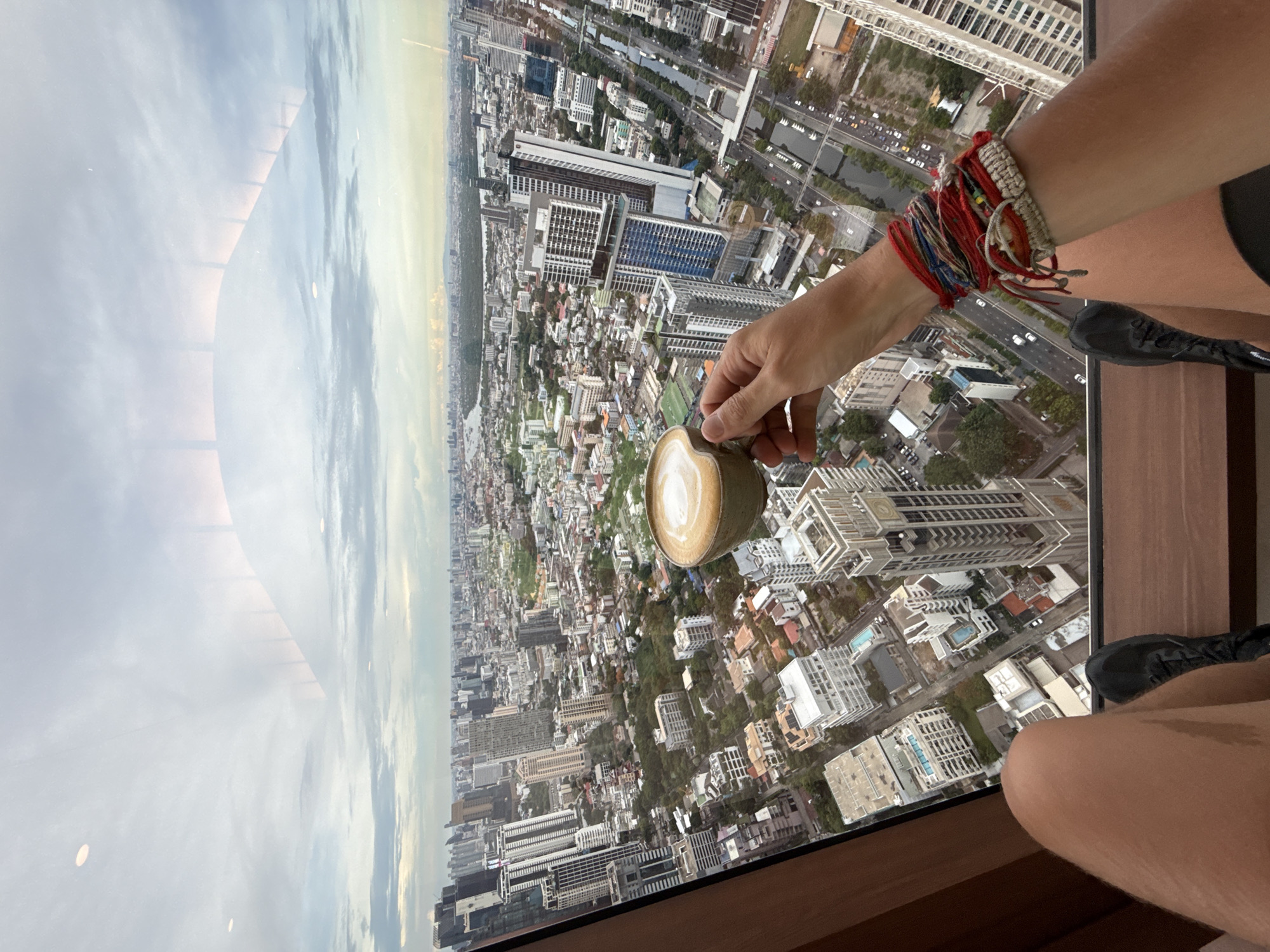 The image shows a high-angle view of the city of Bangkok, Thailand, seen through a window. In the foreground, a person is holding a cup of coffee with latte art. The person is wearing multiple colorful bracelets on their wrist. Their leg and part of their shoe are visible. The background is dominated by the cityscape of Bangkok, including buildings, streets, and patches of green. The sky is visible near the horizon, suggesting the picture was taken during daytime.