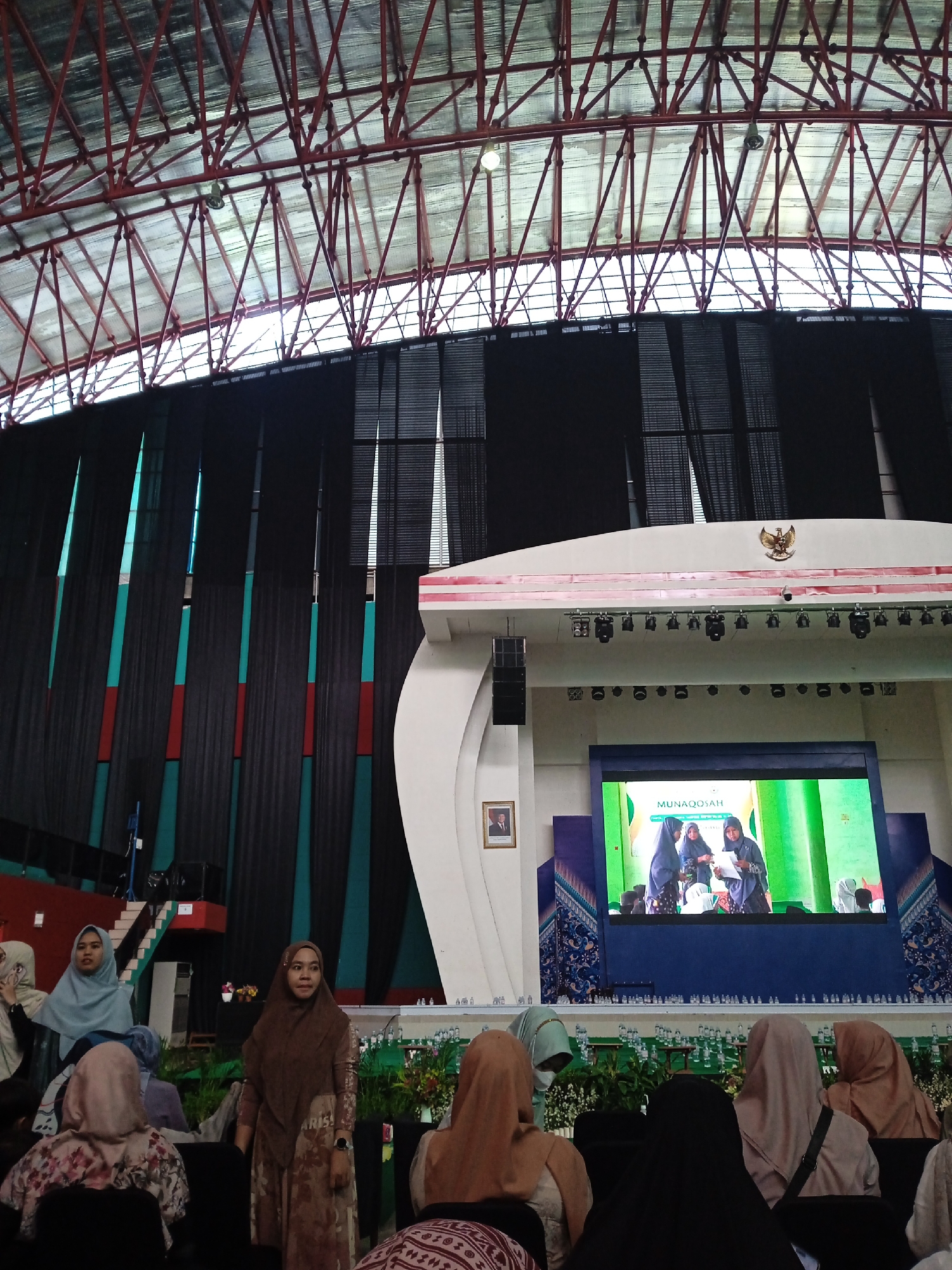 The image shows a presentation stage in what appears to be a large indoor venue, likely a hall or auditorium. The venue's roof structure is visible, featuring a red-painted support system. Black curtains serve as a backdrop to the stage, with intermittent vertical accents of teal and red. A large white architectural element frames the stage, which features a screen displaying an event titled "MUNAQOSAH." The Indonesian coat of arms is affixed to the top of the stage structure.
In the foreground, several women wearing hijabs are seated, presumably as audience members. One woman in a brown hijab is standing in the aisle. The scene suggests an academic or formal event taking place in Indonesia. The city is not discernible from the image itself.