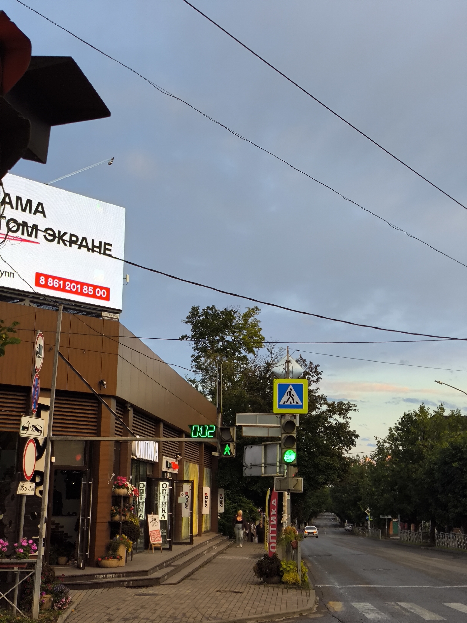 The image depicts a street scene in Yessentuki, Russia, under a partly cloudy sky. On the left, a large billboard displays text in Russian, with a red phone number prominently featured. Adjacent to the billboard is the facade of a building with a wooden-paneled exterior, featuring multiple storefronts. Signage in Russian, including "ОПТИКА" (Optics), is visible on the shops. A pedestrian crossing is marked on the road, and a traffic light shows a green pedestrian signal. Several trees line the right side of the street, which recedes into the distance. A few cars are present on the road, and a person is walking on the sidewalk. The overall scene suggests an urban environment during daylight hours, possibly late afternoon or early evening, given the lighting.