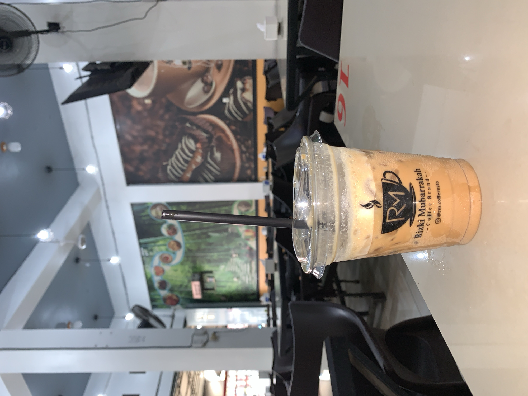 An eye-level, medium shot captures a plastic cup of iced coffee on a white table in what appears to be a restaurant in Tambonbaroh, Indonesia. The cup is mostly full of a light brown, frothy liquid with ice cubes. The cup has a clear plastic lid and a black straw. A logo on the cup reads "RM Rizki Mubarakah - Coffee Brand -" and includes an Instagram handle "@rm.coffeeresto". The background shows a brightly lit interior with dark tables and chairs. Large murals are visible on the walls: one depicts a forest scene with circular elements, and another shows coffee cups and pastries. The ceiling is blue and white, with hanging lights. A ceiling fan is visible in the upper right corner. The scene is static and appears to be taken during daylight hours.