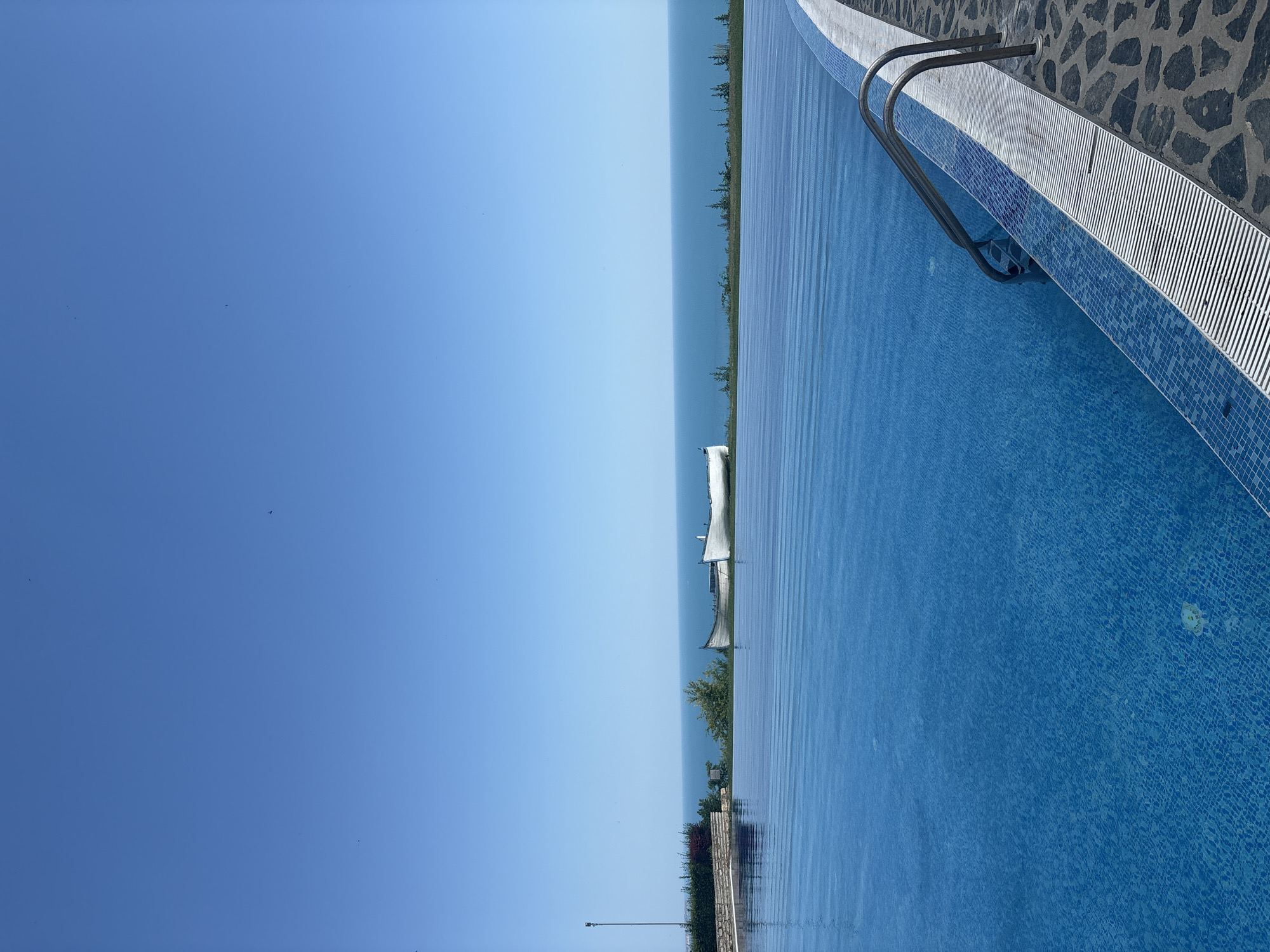An infinity pool overlooks a calm body of water, likely a sea or large lake, under a clear blue sky. The pool's edge is tiled with small blue mosaics, and a metal ladder dips into the water. The surrounding deck is made of dark, irregularly shaped stones. In the distance, a strip of green vegetation, including trees, separates the pool from the water. There are no people visible in the image, suggesting a peaceful and unoccupied setting. The lighting indicates it is daytime, possibly late morning or afternoon, with bright sunlight casting subtle reflections on the water's surface. The overall impression is one of tranquility and a serene vacation setting.