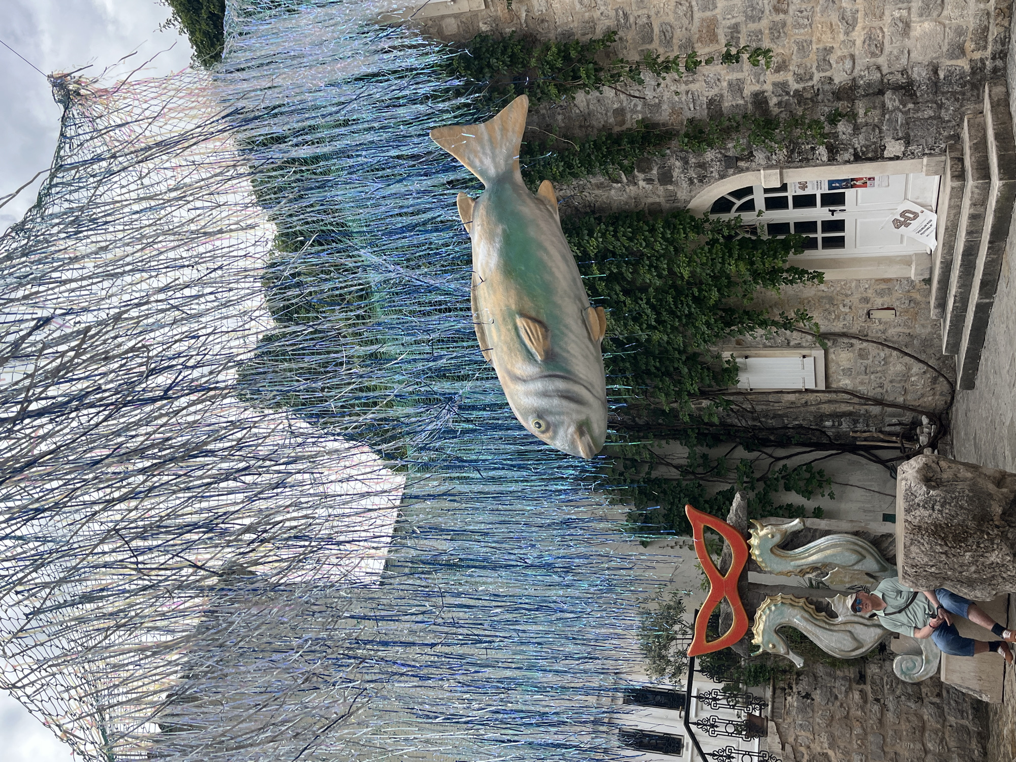 The image shows an outdoor setting, likely a street or public area in a city. The primary focus is a large, decorative fish hanging in the air. Above the fish is a net-like structure filled with silver and blue streamers. Behind the fish is a stone building with ivy growing on the wall. The building has a white door with windows, and steps leading up to the door. To the right of the fish, there's a sculpture with two seahorses and an orange mask. A person wearing sunglasses and casual clothes is sitting on a stone structure next to the seahorse sculpture. The image appears to be taken in a city, but the specific city is not identifiable from the image.