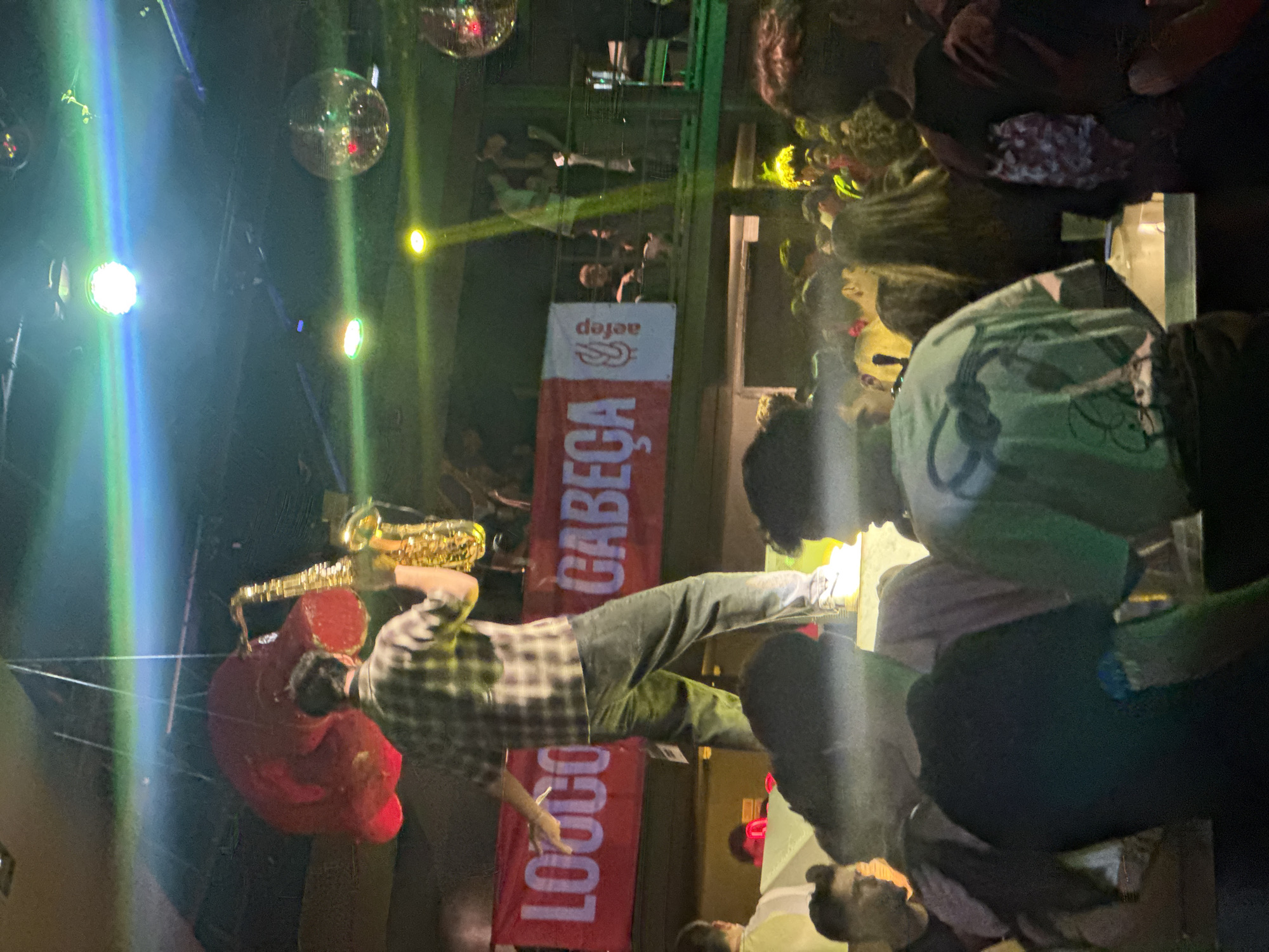 A person playing a saxophone while sitting in a red chair is at the club named "CABEÇA" in the city of Porto, Portugal. The club is packed with people and there are disco balls hanging from the ceiling. A sign of the club is seen hanging in the background.