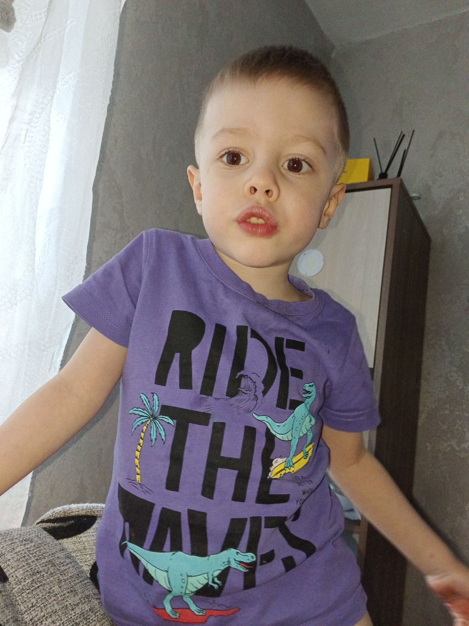A young boy is standing indoors. He has short, light brown hair and brown eyes. He is wearing a purple t-shirt with a large graphic that says "Ride The Waves," featuring a palm tree and dinosaurs. He has a slightly open mouth. The background shows a white curtain, a gray wall, and a piece of furniture with a yellow box on top and some black sticks in a container. A piece of grey fabric is in the bottom left corner. There is no other information provided that relates to the location in the city of Chaikovsky, Russia.