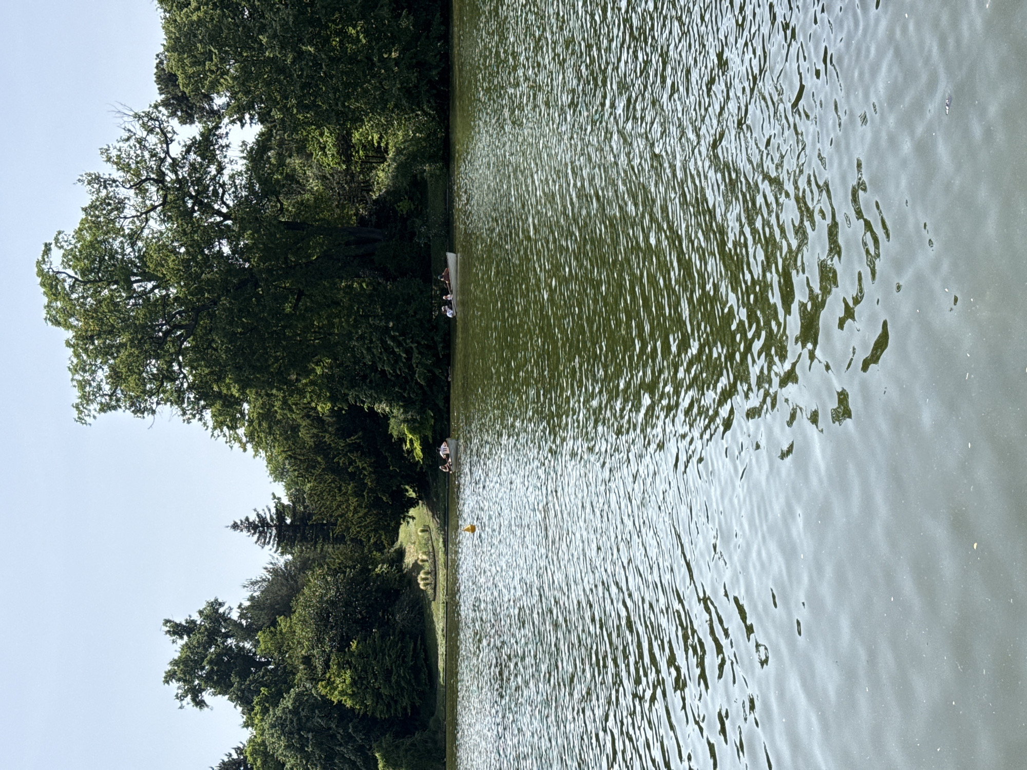 A scenic view of a lake or pond, possibly in a park, with trees lining one side and sunlight shimmering on the water's surface. Two people are sitting on a bench at the water's edge. A yellow buoy floats in the water. It could be located in one of the many parks and gardens in Paris, France.