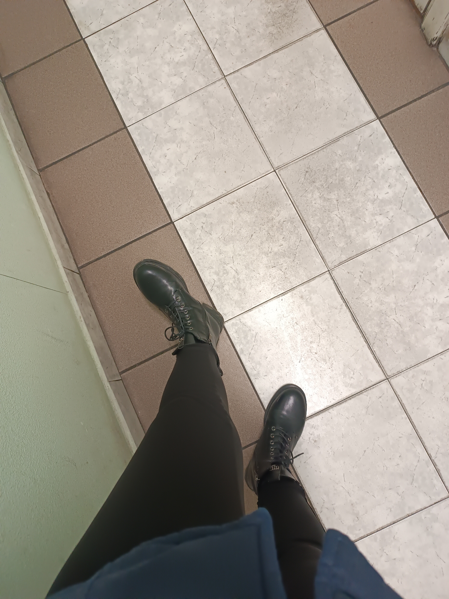 The image shows a person standing on a tiled floor. The tiles are arranged in a grid pattern with alternating rows of light beige and brown tiles. The person is wearing black leggings and black lace-up boots. The person is also wearing a blue jacket, with only the bottom edge visible. The image is taken from a first-person perspective, looking down at the person's legs and feet. Based on the location clues, this scene may be inside a building in Chaikovsky, Russia, likely a public building given the type of tiled floor and wall.