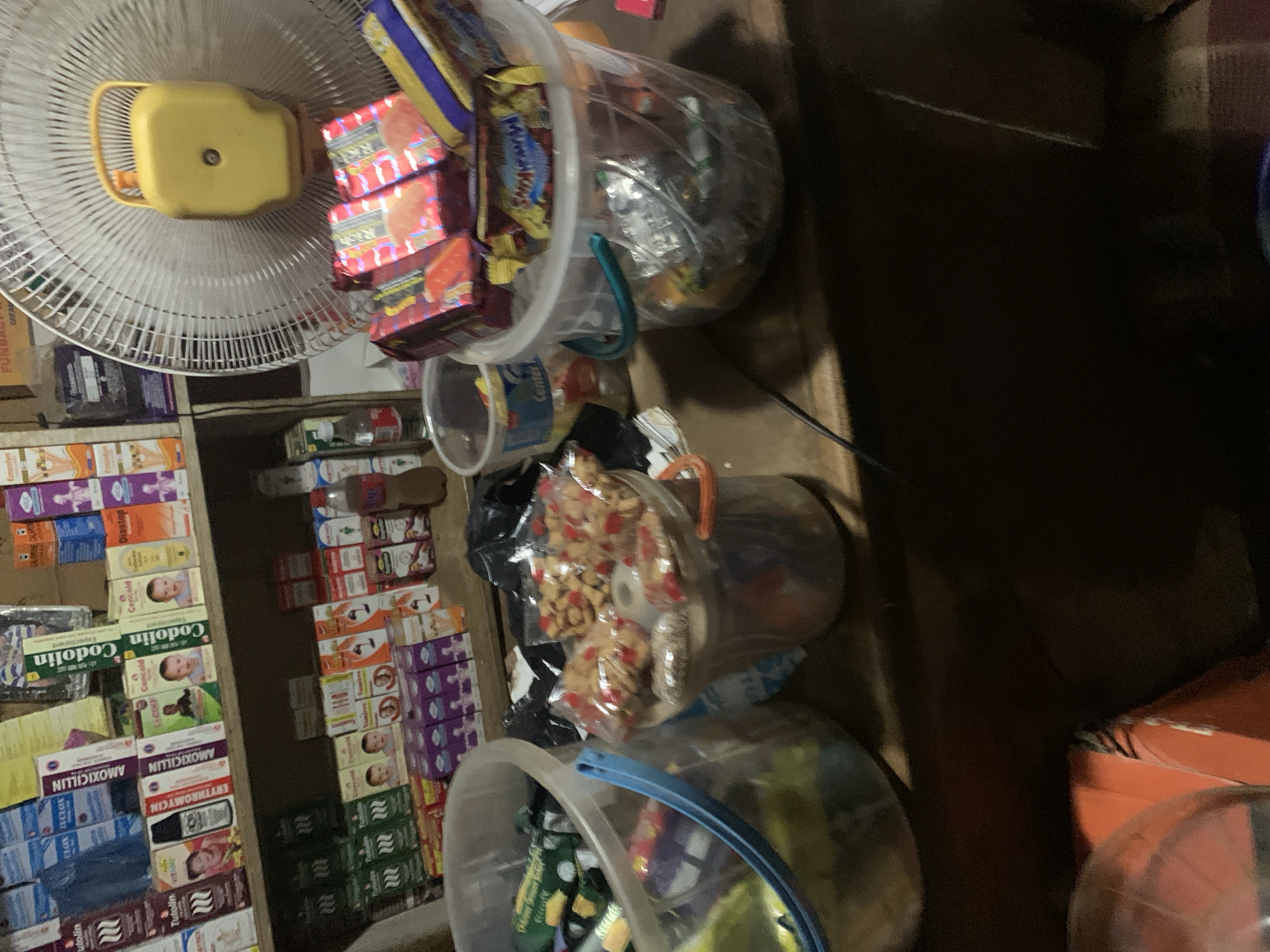 The image shows the interior of what appears to be a small store, possibly in Gbeleko, Nigeria, given the medications labeled in English and the context of the prompt. A shelf on the left side displays boxes of various medications including "Codolin" and "AMOXICILLIN", stacked vertically. In front of the shelf are three transparent plastic buckets. The topmost bucket contains what looks like sweets or candy. The middle bucket appears to contain bags of peanuts and other small items. The bottom bucket is also filled with assorted items, including wrapped goods, appearing to be snack items. A yellow and white electric fan is positioned above the merchandise, likely to provide ventilation. The background features a dark wooden structure.