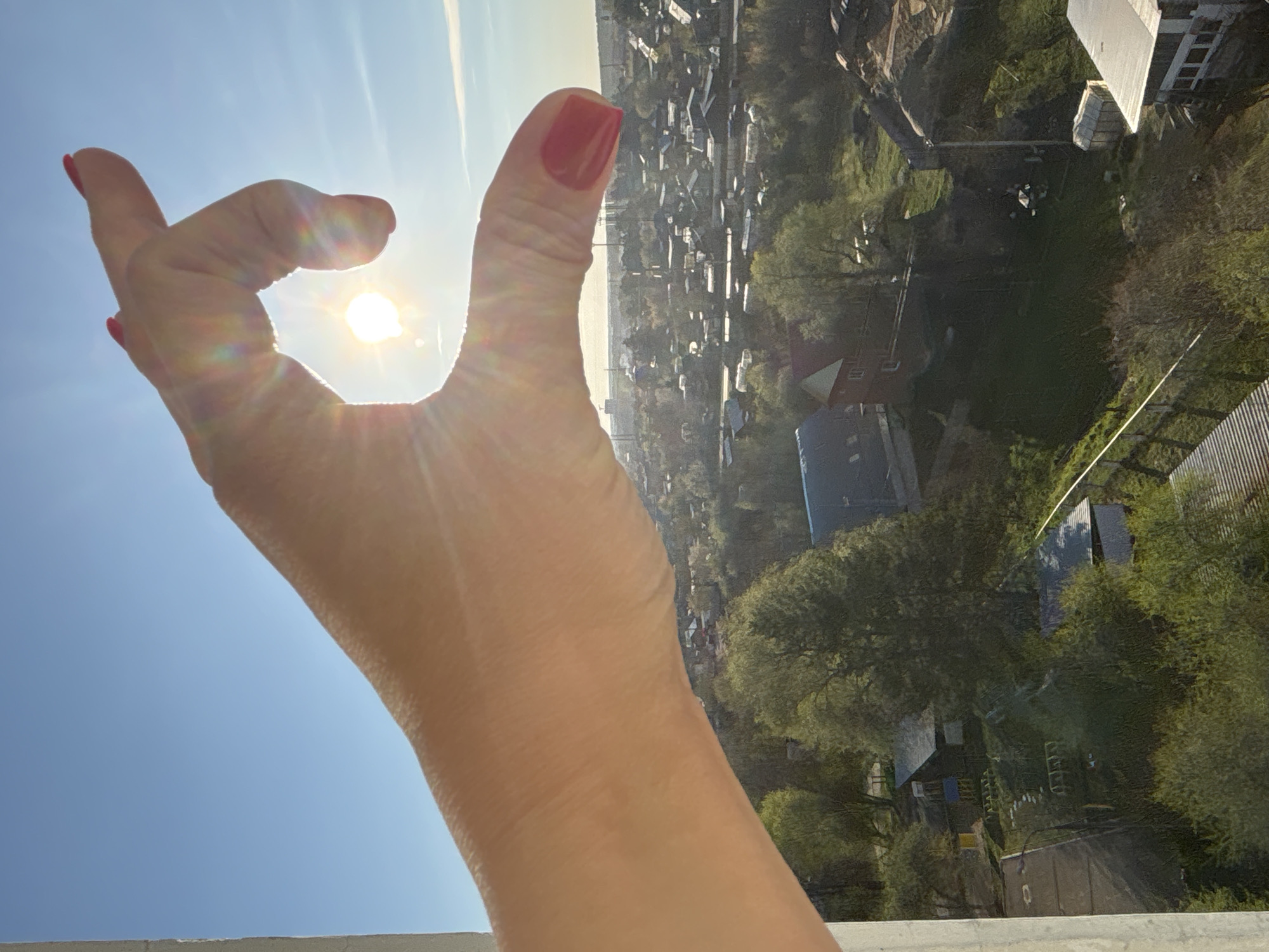 A woman's hand with red nail polish is positioned against a bright, sunny sky. The sun shines through the opening formed by her fingers. In the background, a view of the city of Chaikovsky, Russia, is visible. Buildings, trees, and streets form the urban landscape.