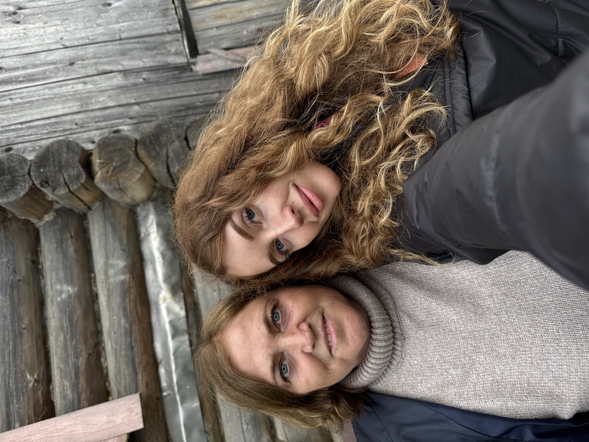 A close-up, vertically oriented portrait features two women posing together, likely in a selfie, against a rustic wooden background. Both subjects are looking directly at the camera with pleasant, relaxed expressions.
The woman positioned higher in the frame appears younger, with long, voluminous, reddish-blonde curly hair that frames her face. She has striking blue eyes and a subtle smile, with her tongue peeking out slightly. She is dressed in a dark, possibly black, puffy jacket.
Below her, nestled closely, is an older woman with shoulder-length, light brown hair and blue eyes. She is smiling gently. She wears a light gray, ribbed turtleneck sweater beneath a dark jacket, similar in style to the younger woman's.
The background provides strong contextual cues, featuring weathered wooden structures consistent with a traditional rural setting in Maksimovo, Russia. To the left, a wall constructed from horizontal, round-cut wooden logs, characteristic of a log cabin, is prominent. Further back and to the right, a section of vertical wooden planks can be seen. In the lower right quadrant, a piece of corrugated metal siding or roofing is visible, along with another wooden plank. The lighting is soft and even, suggesting an overcast daytime, and the warm clothing worn by both women indicates cool weather. There is no visible text in the image.