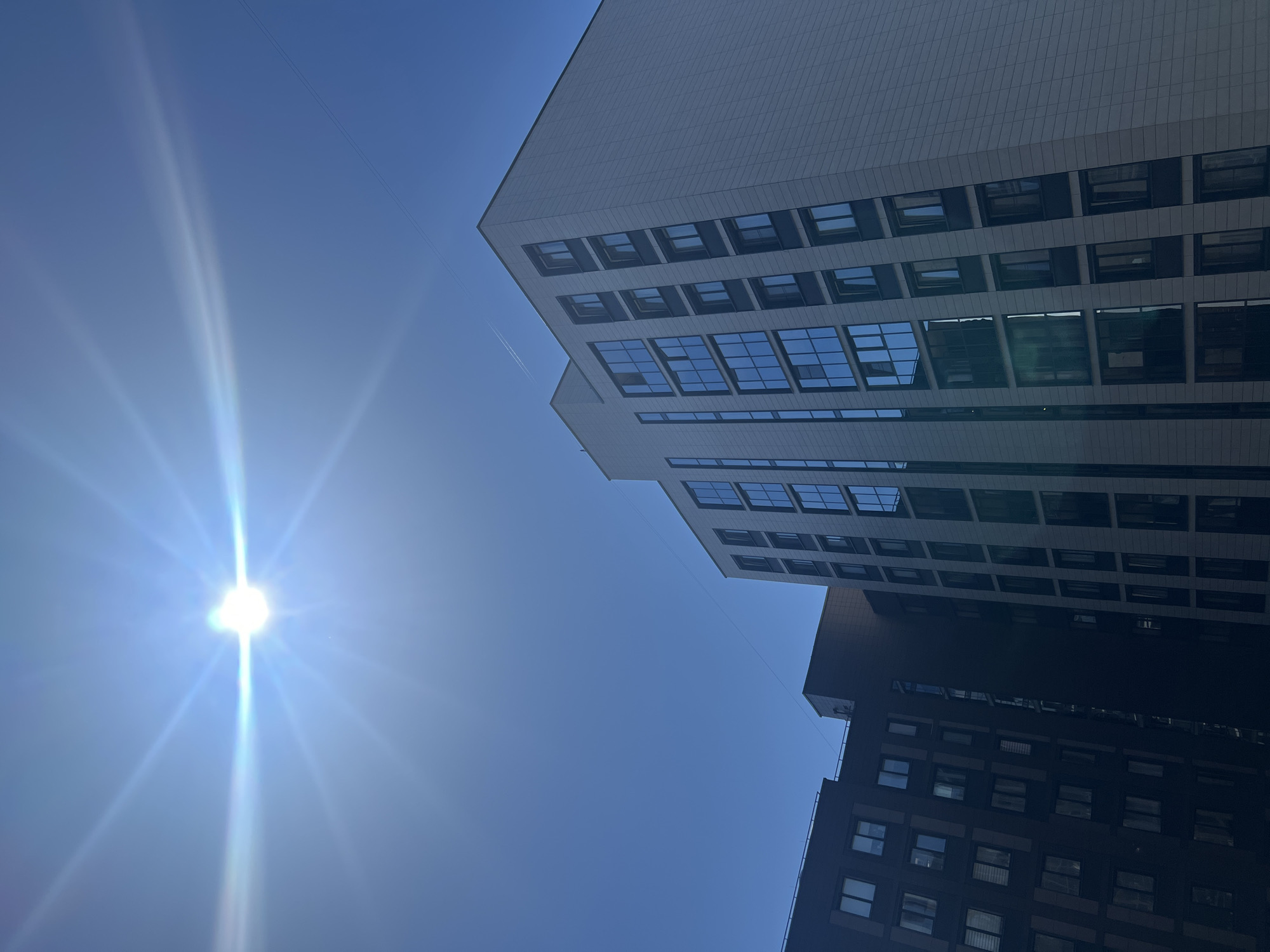 The image depicts an urban scene in the city of Saint Petersburg, Russia, featuring two modern high-rise buildings under a clear, bright blue sky. The sun, positioned in the upper-left quadrant, is a brilliant white light source generating a pronounced starburst lens flare with prominent horizontal light streaks. A faint white aircraft contrail is discernible in the upper-right area of the sky.
The primary building on the right is a multi-story structure with a light gray or off-white facade composed of rectangular panels. Its multiple windows, arranged vertically, exhibit a reflective blue tint where illuminated by the sun, while windows on its shadowed side or facing away from the sun appear dark. The building on the left is partially visible, presenting a darker, possibly brown or dark gray facade, also featuring numerous windows. Both structures suggest contemporary residential or commercial architecture. Thin utility wires traverse the upper portion of the frame: one extends diagonally from the upper-center to the upper-right, and another, less distinct wire connects the upper sections of the two buildings. No people or specific identifying landmarks within Saint Petersburg are visible. The scene is illuminated by strong daylight.