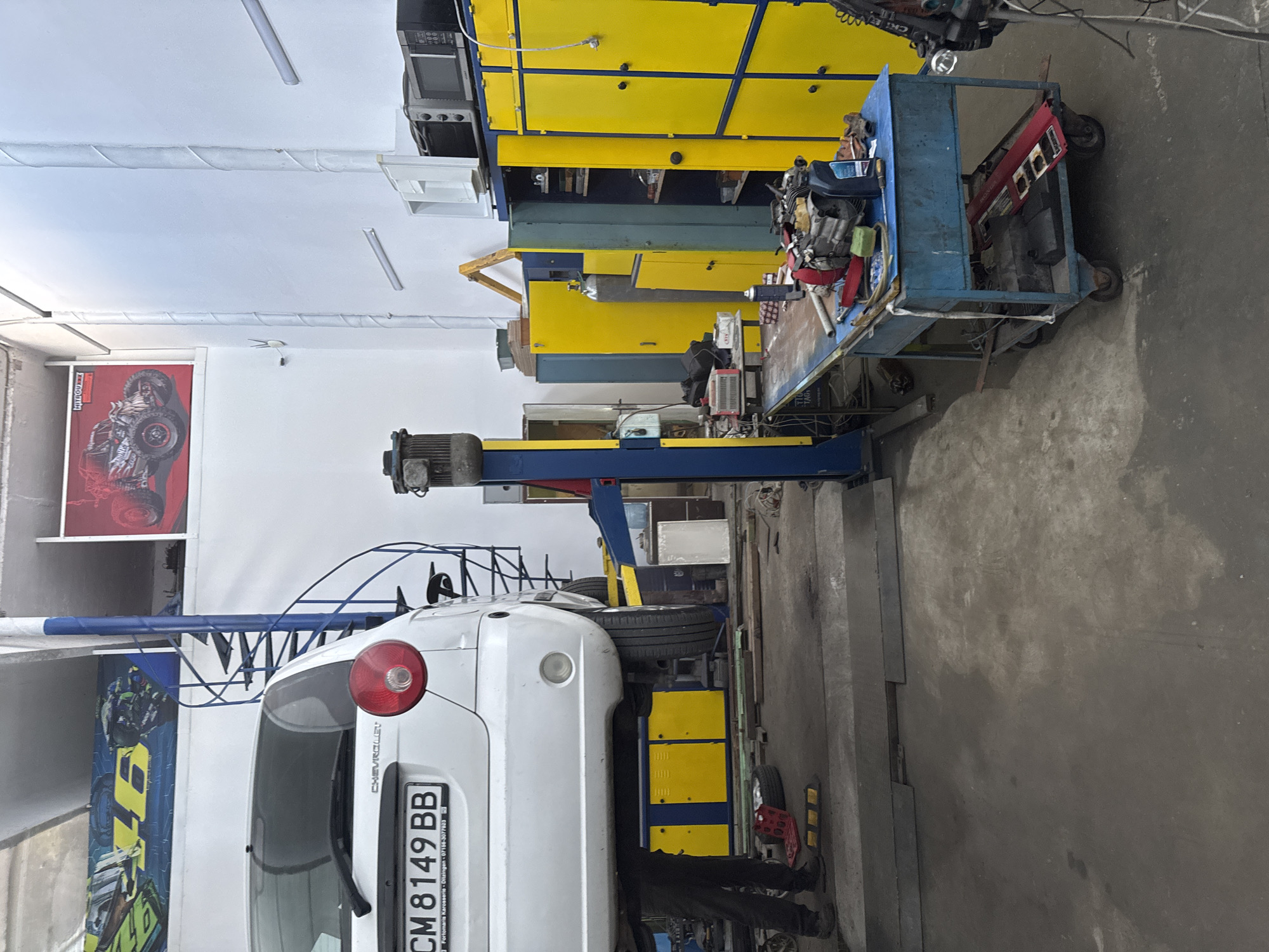 The image depicts the interior of a car repair workshop in Smolyan, Bulgaria. A white Chevrolet car is elevated on a hydraulic lift. The workshop is equipped with various tools and equipment, including a workbench with tools, yellow storage cabinets, a welding machine, and a gas cylinder. A person, dressed in dark pants and shoes, is standing near the car. The workshop appears to be well-lit, with fluorescent lighting and natural light entering from the windows. The walls are painted white, and the floor is concrete. A spiral staircase is visible in the background, leading to an upper level. There are posters on the wall, including one with a racing theme. The overall scene suggests an active and functional car repair facility.