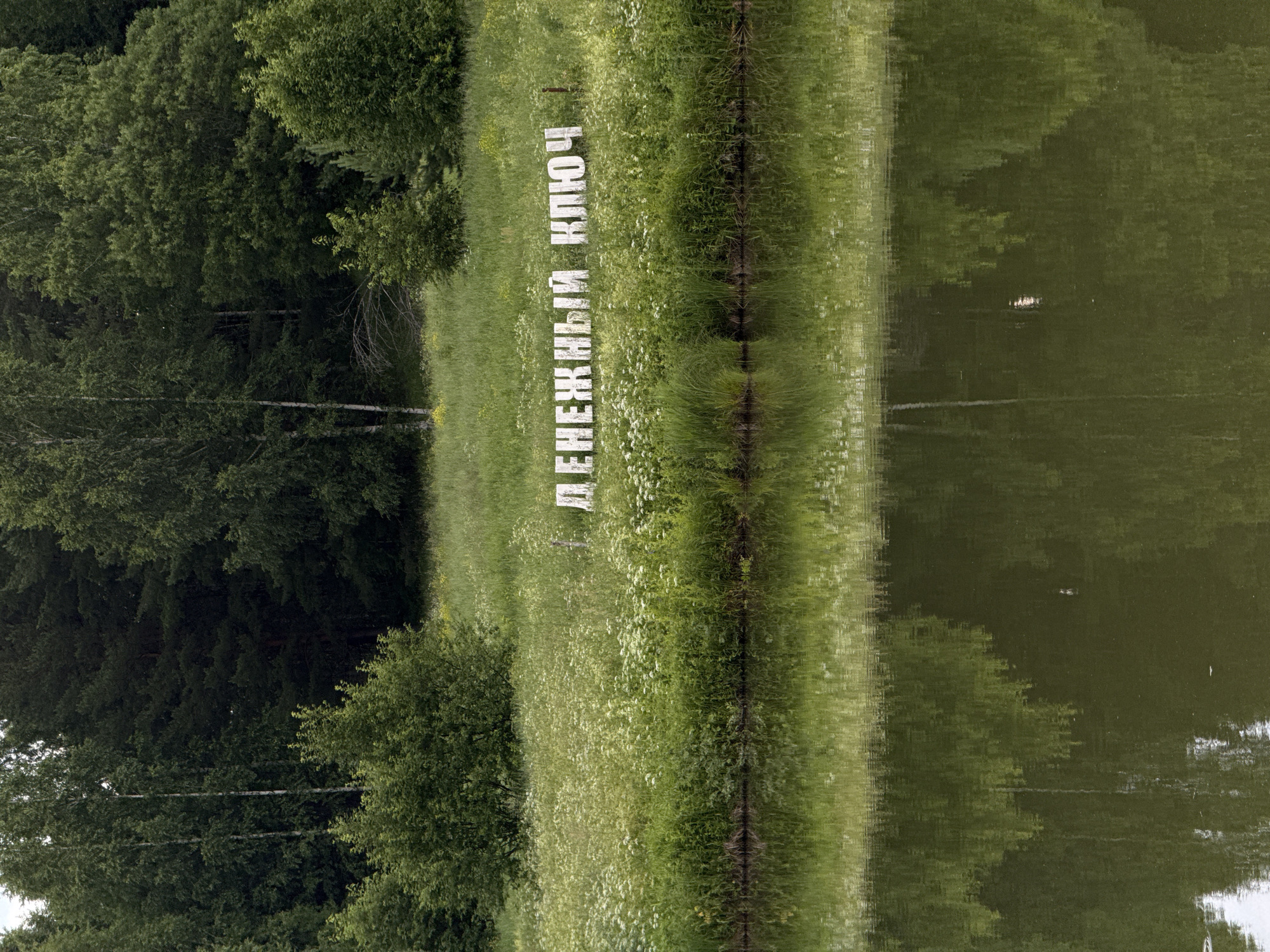 The image shows a reflective body of water in the foreground with a grassy embankment and a dense forest in the background. White, block-style letters spelling "ДЕНЕЖНЫЙ КЛЮЧ" (which translates to "Money Key" in Russian) are placed on the grassy embankment. The letters appear to be made of wood or a similar material and are spaced apart. The environment is lush and green, indicative of a natural setting. The water's surface shows a clear reflection of the embankment and trees. This scene is located at the Votkinsk Reservoir in Votkinsk, Russia. There are no people or animals visible in the image.