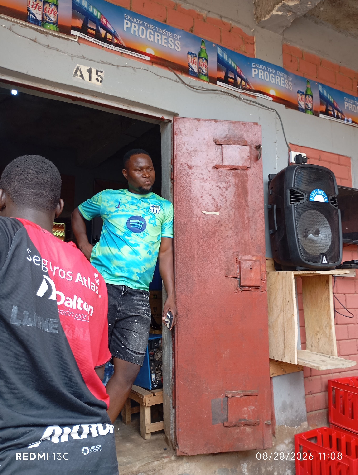 A medium shot captures two men standing near the entrance of a business establishment in Shendam, Nigeria. The man on the left, seen from behind, wears a black shirt with red accents and the text "Seguros Atlas Dalton" and "LAME Z MA" printed on the back. He also has "REDMI 13C" and "ORLEGI SPORTS" visible near the bottom. The man on the right, facing the viewer's left, is wearing a tie-dye t-shirt with blue and green swirls, and dark, distressed jeans. He has his left hand on his hip and is holding a dark object in his right hand.
The background reveals a rustic storefront. A large, weathered red metal door dominates the right side of the frame, featuring a rectangular panel with a small keyhole. To the left of the door, a sign with "A15" is affixed to a grey surface. Above the entrance, a banner displays "ENJOY THE TASTE OF PROGRESS" with images of beer cans and bottles. A black speaker sits on a wooden shelf to the right of the red door, positioned in front of a television. Red plastic crates are stacked to the far right. The overall environment appears to be an outdoor market or shop front, with brick walls and a concrete floor. A timestamp "08/28/2026 11:08" is present in the bottom right corner.