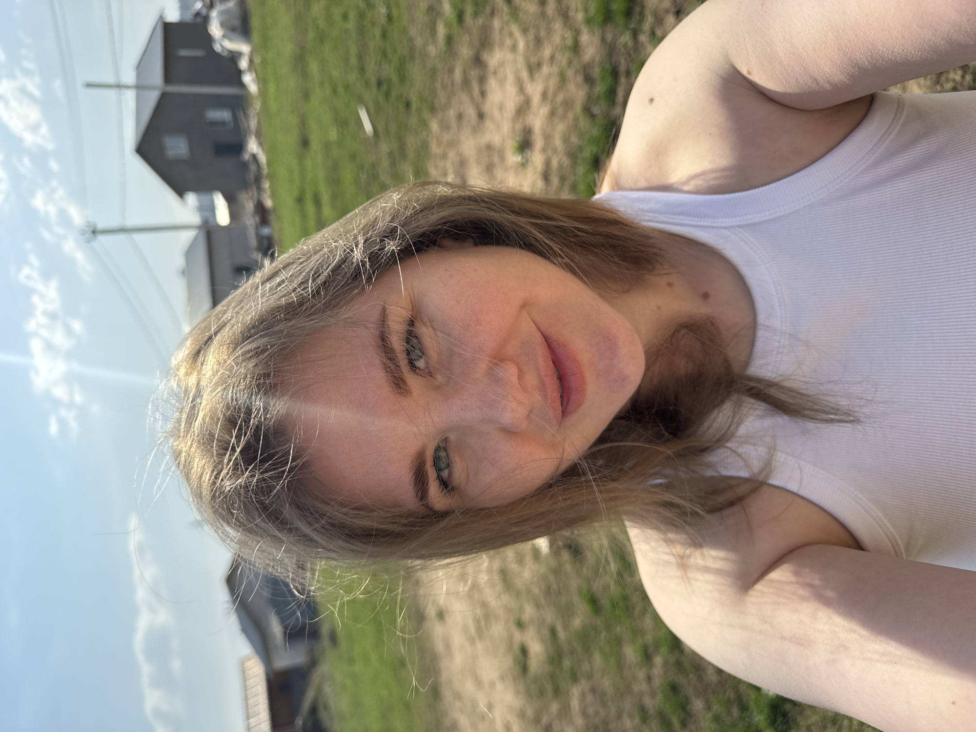 A selfie taken outdoors in the city of Ферма, Russia, shows a woman with fair skin, blonde hair, and light-colored eyes wearing a white tank top. The background includes a grassy area, a grey building, and utility poles against a blue sky. The woman is smiling and looking directly at the camera.