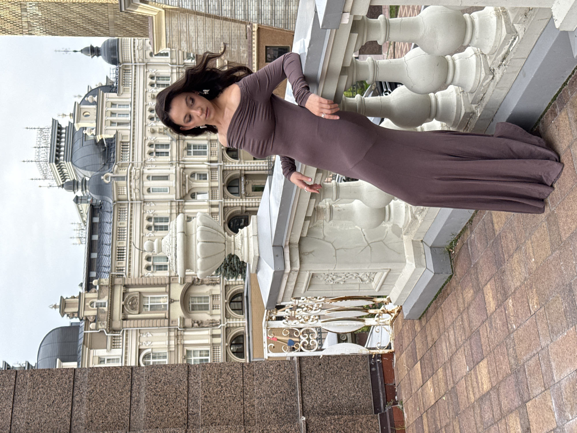 A woman in a long, off-the-shoulder, brown dress leans against a white ornate railing in an outdoor urban setting. Her hair is dark and flows around her face. She is wearing earrings and rings. The railing overlooks a cobblestone patio and a street with a car. Behind her, a large, multi-story building with intricate architecture and domes, characteristic of Kazan, Russia, stands against a cloudy sky. The building features ornate detailing, arched windows, and balconies. To the left of the frame, a section of a granite-clad building is visible. The overall scene suggests a view from an elevated position in the city.