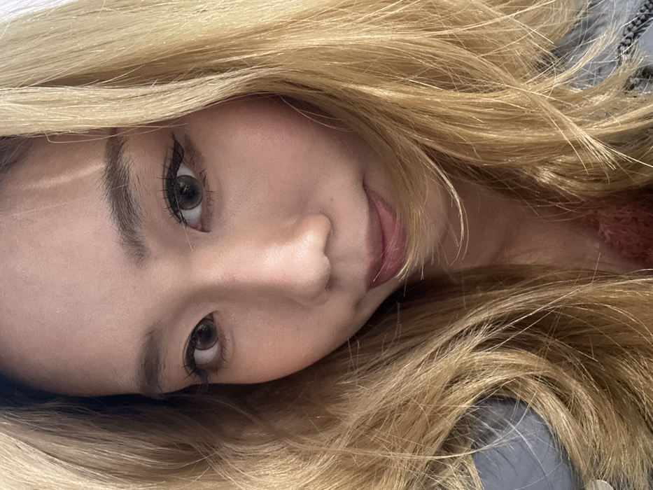 A close-up portrait shows the upper torso and head of an individual with light skin and East Asian features. The individual has long, wavy, bleached blonde hair that frames the face and partially covers the shoulders. The eyes have dark brown irises, possibly enhanced by colored contact lenses, and are lined with dark eyeliner, winged at the outer corners. Eyelashes are prominent. Eyebrows are dark brown and well-defined. The nose is visible, and the lips are full with a light pinkish-brown tint. A slight shadow is present on the left side of the face. A light blue garment is visible on the left side of the frame, underneath the hair, and a pink, textured garment is visible on the lower right. The individual's gaze is directed towards the camera with a neutral expression. The background is indistinct due to the tight cropping. The location in Shapingba District, China, cannot be determined from the visual information present in the image.