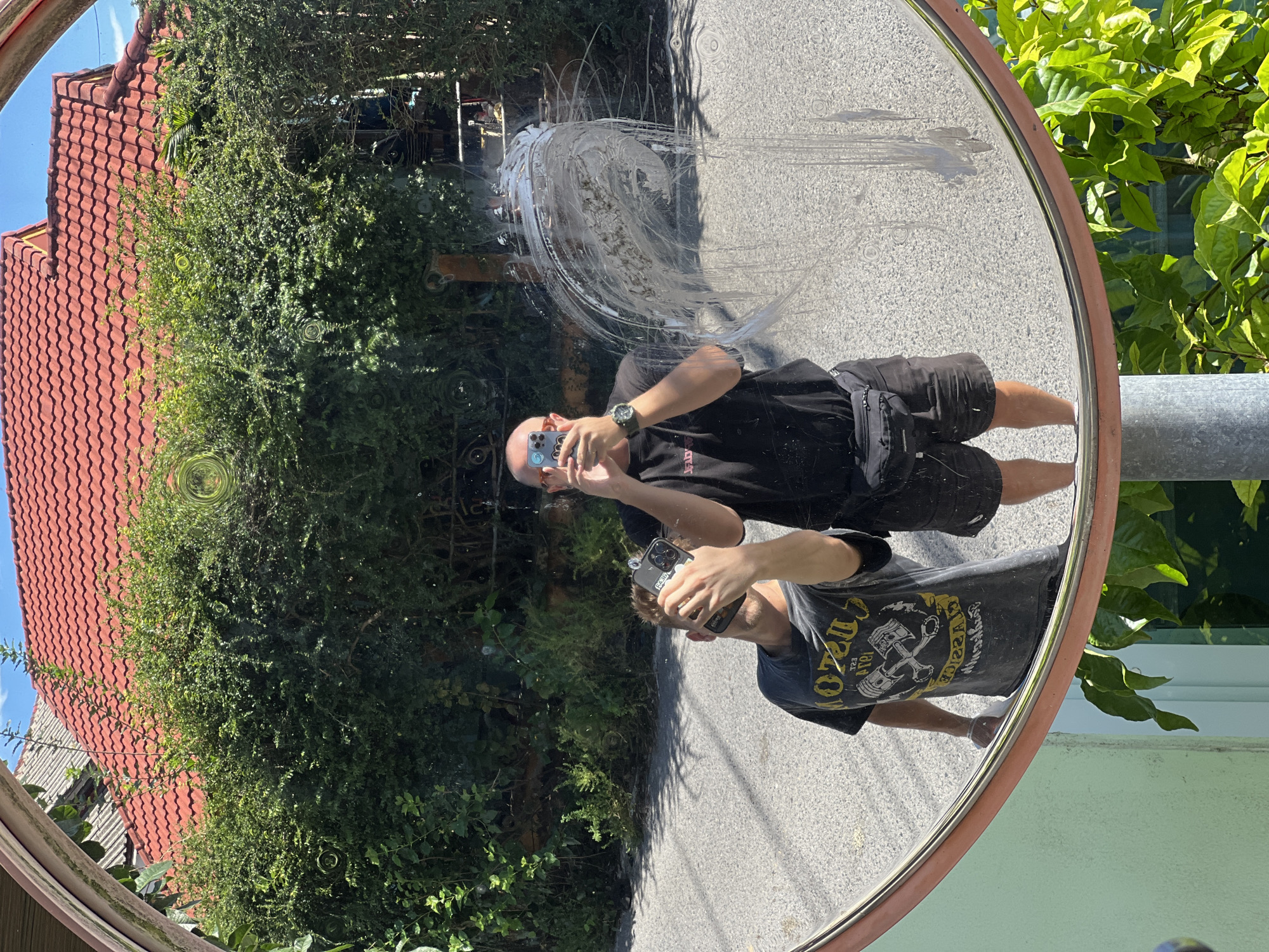 A man wearing a grey t-shirt with a graphic print, black shorts, and a black fanny pack takes a selfie with his phone in a convex mirror. He is standing to the left of another man, who is wearing a black t-shirt and black shorts and is also taking a selfie with his phone. The mirror reflects a street scene with a red-tiled roof house and green bushes. The mirror has some smudges and streaks on its surface. The ground is paved with asphalt. The lighting suggests it is daytime and sunny.