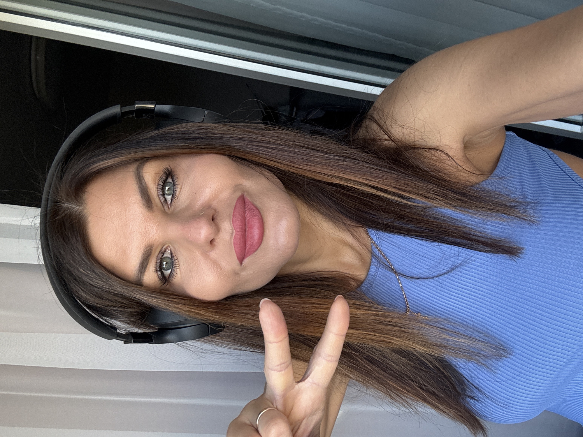 A woman is depicted indoors with a window behind her. She is wearing headphones and a blue top, holding up a peace sign with her hand. She has brown hair, grey eyes, and is wearing lipstick. The location cannot be determined from the image.