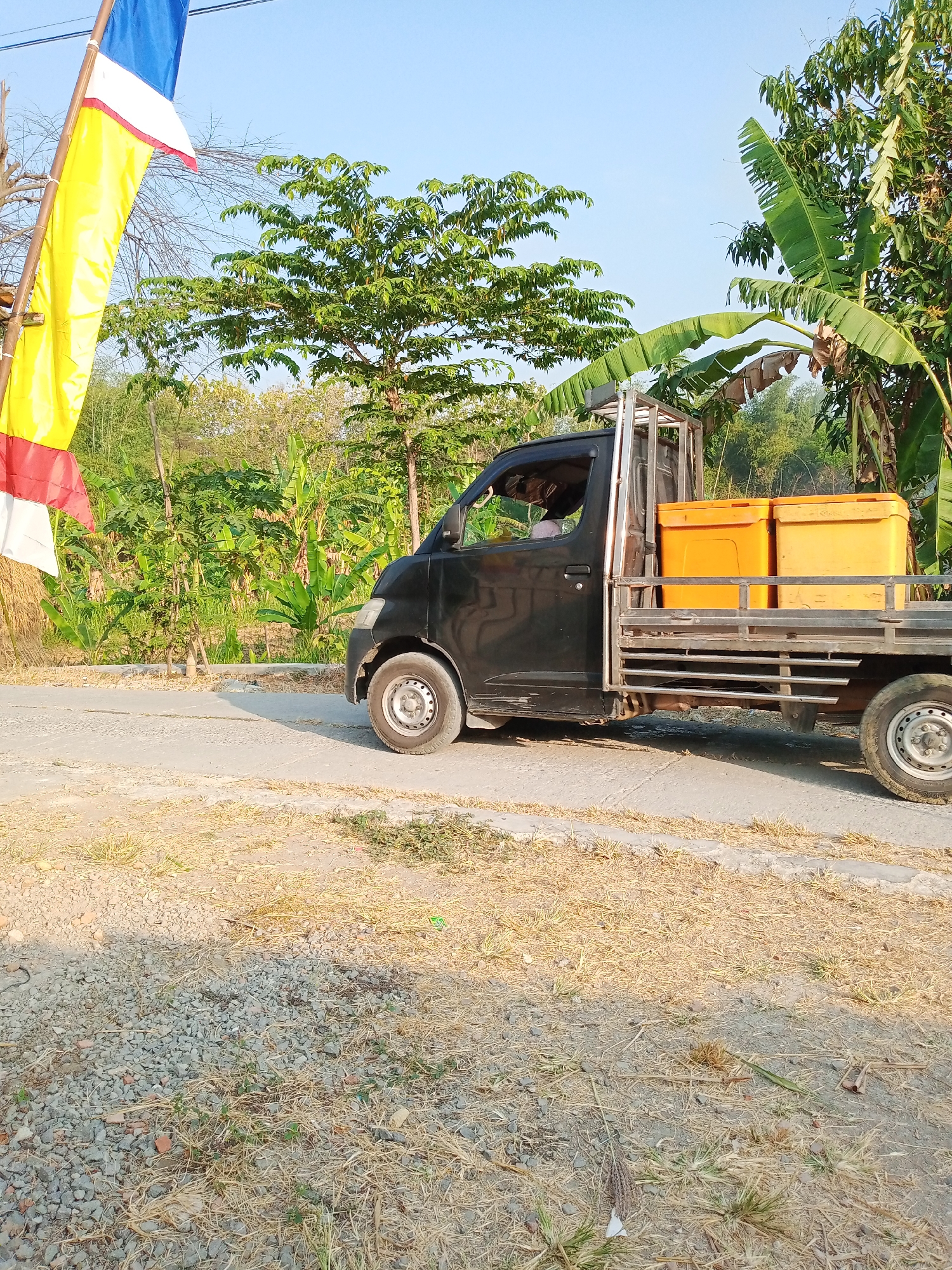 An outdoor, eye-level, full shot captures a black utility truck parked on a dirt and asphalt road in the city of Kemiri, Indonesia. The truck, with its bed carrying two large orange containers, is positioned in the center-right of the frame. Behind the truck, a dense green tropical foliage of trees and banana plants lines the background. A flag with red, white, and yellow stripes is visible in the upper-left corner of the image. The sky is a clear, light blue. The foreground is mostly dry grass and gravel.